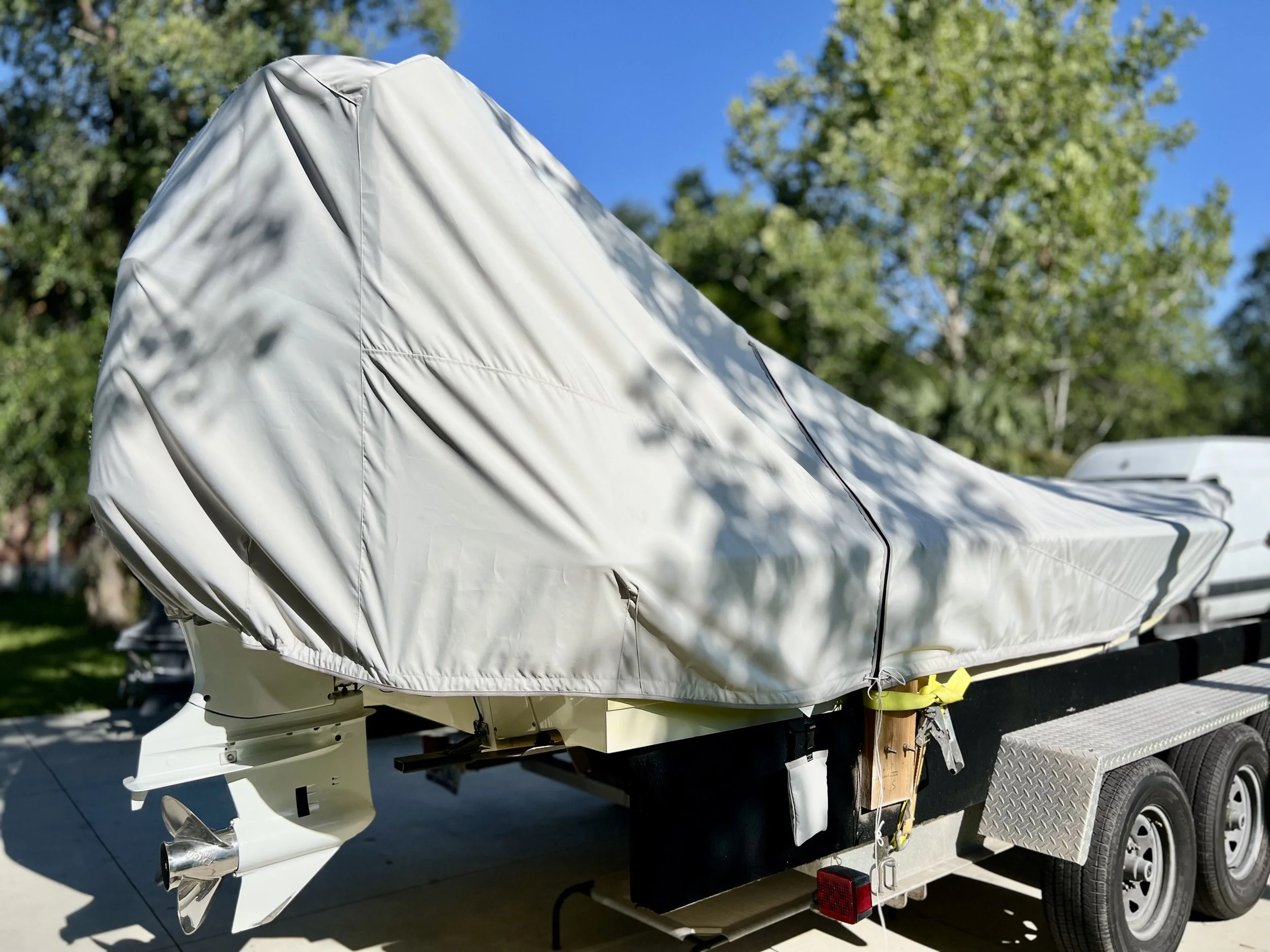 A boat on a trailer covered with a white protective cover, parked outdoors under trees with a clear blue sky.