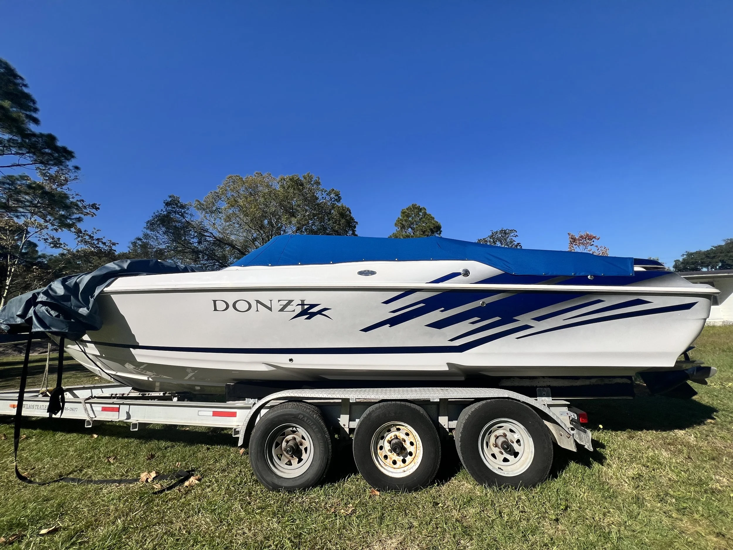 A white speedboat named DONZI on a triple-axle trailer, covered with a blue tarp, on a grassy area under a clear blue sky with trees in the background.