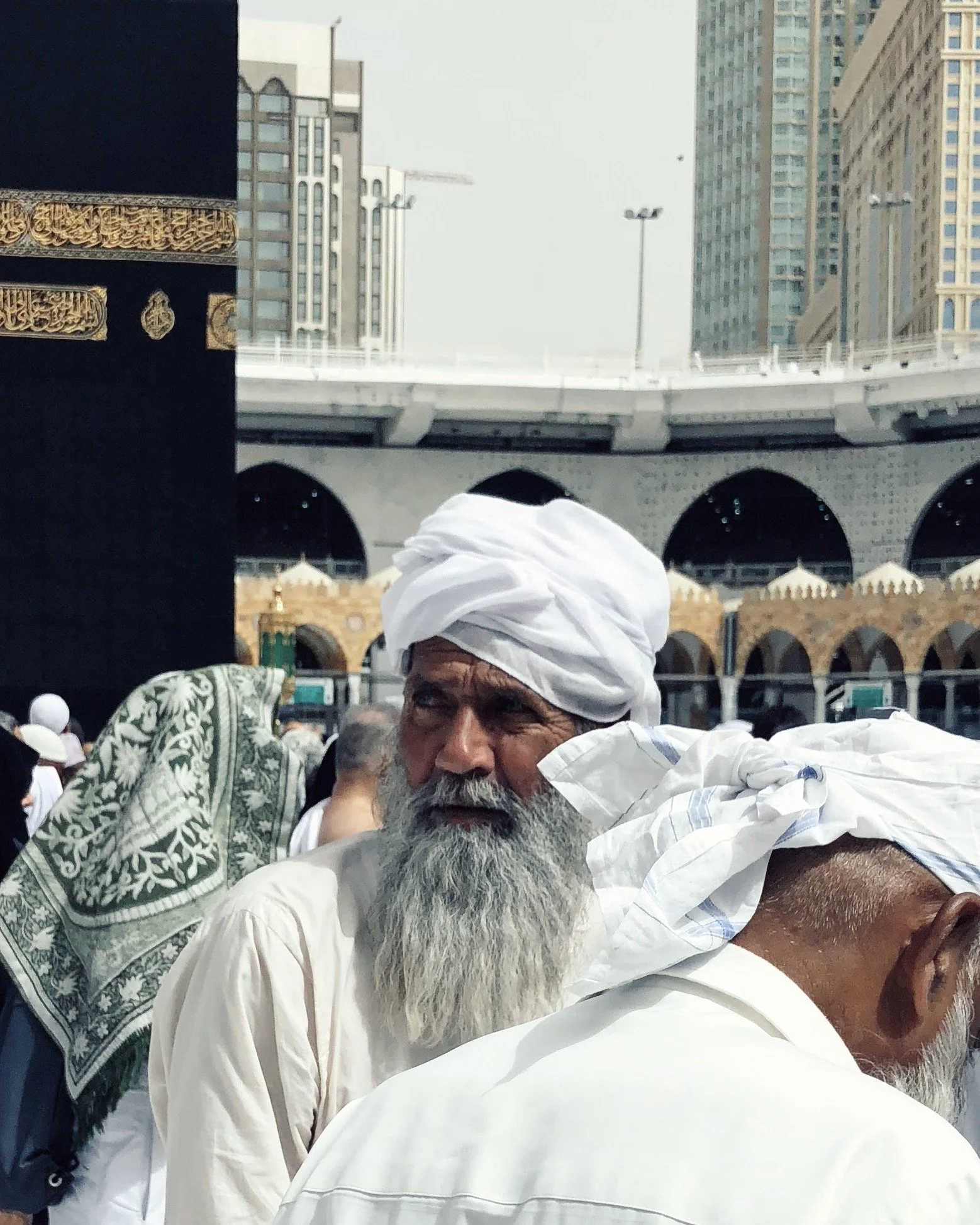 A crowd of people, including an elderly man with a white beard and traditional white turban, gathered outdoors near a mosque with a black and gold architectural structure, with tall buildings in the background.