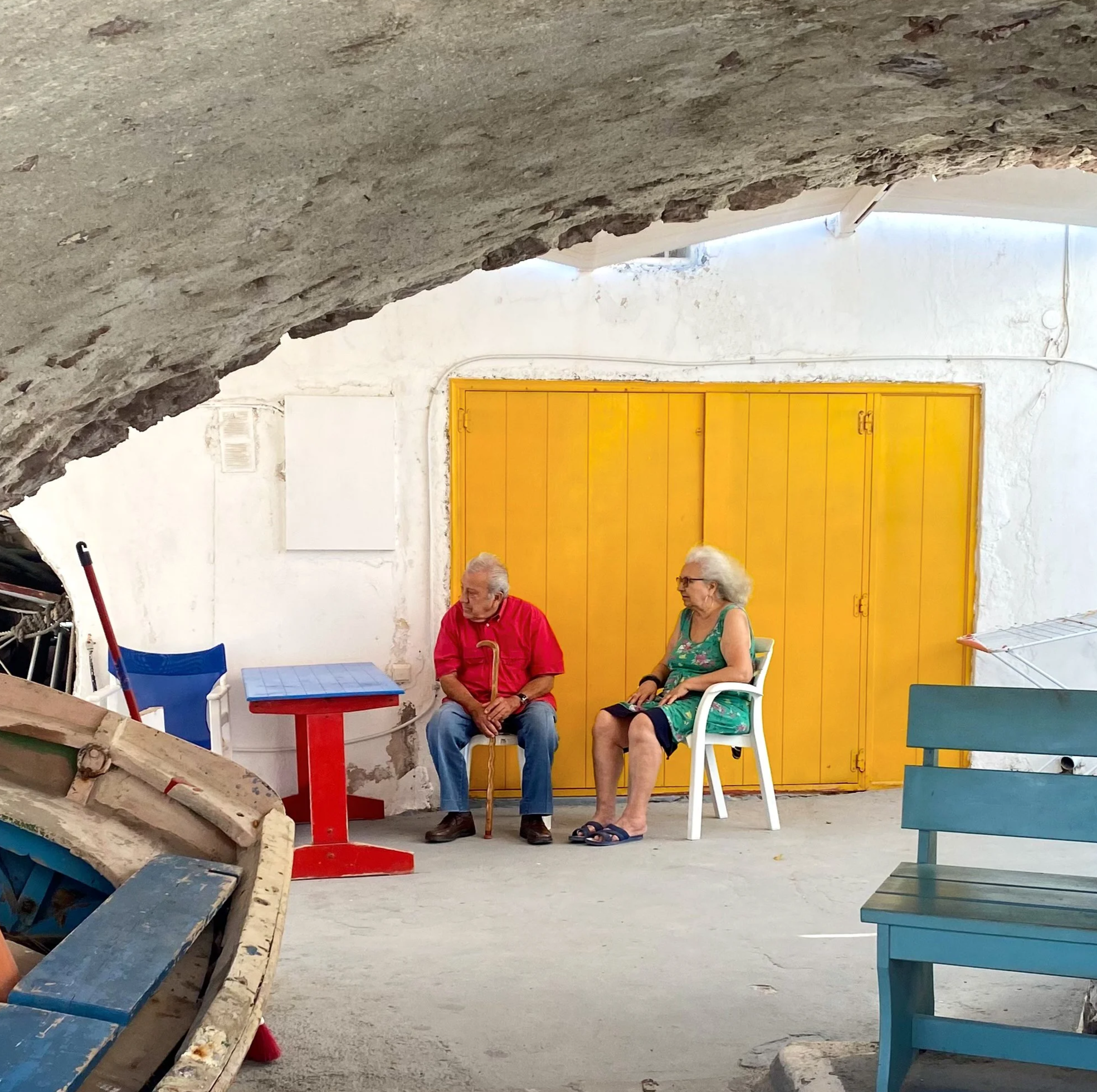 Two elderly people sitting in a shaded area, a man with a cane in a red shirt and an elderly woman in a green dress with glasses, sitting on white plastic chairs, near a yellow door and a small blue and red table.