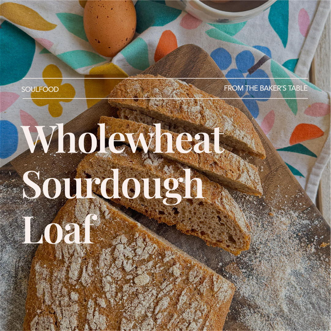 A loaf of whole wheat sourdough bread, partially sliced, on a wooden cutting board. In the background, there are eggs, a cup of coffee, and colorful floral cloth.