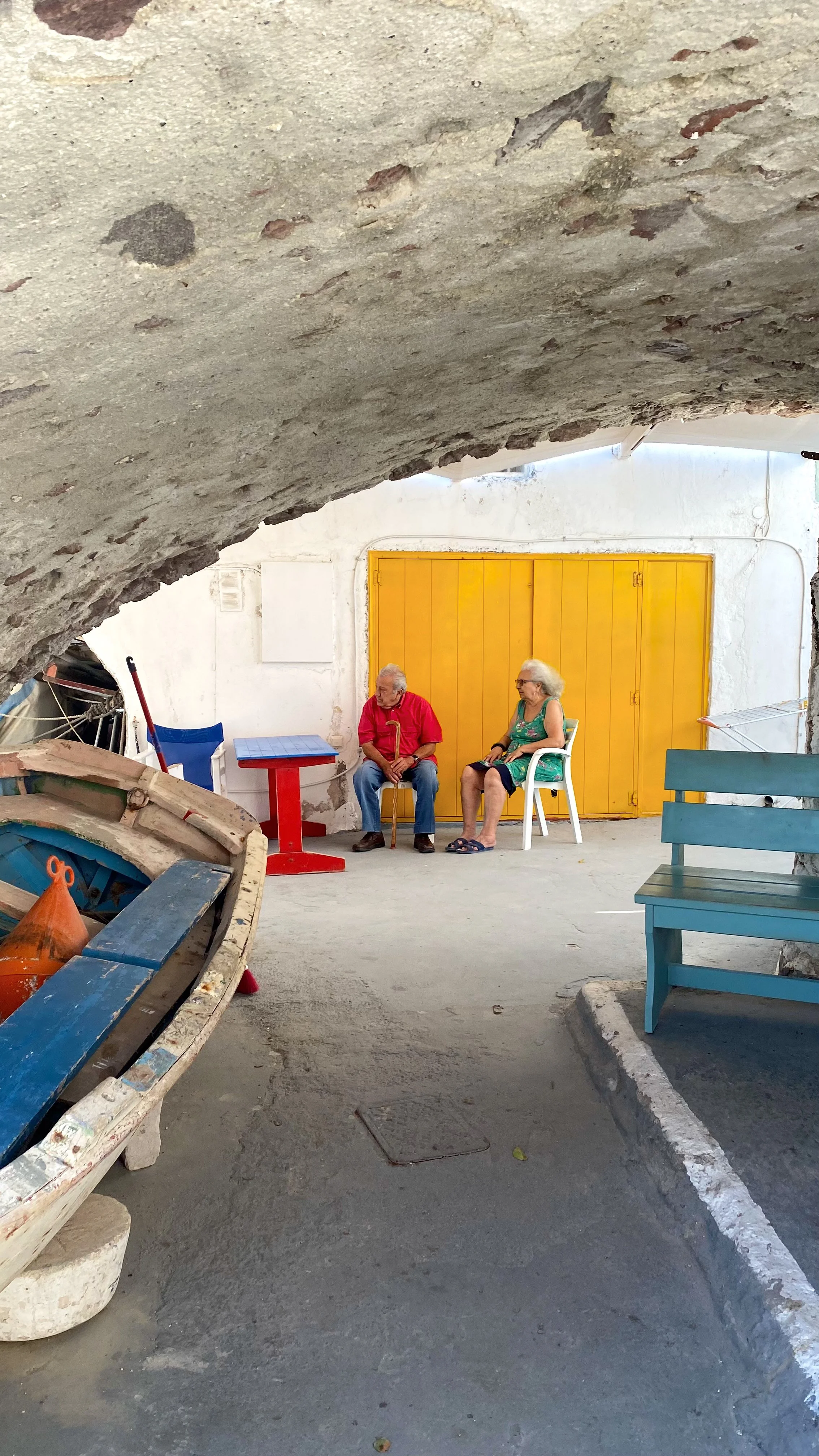 Two elderly people sitting on chairs in a shaded outdoor area with a yellow door in the background.