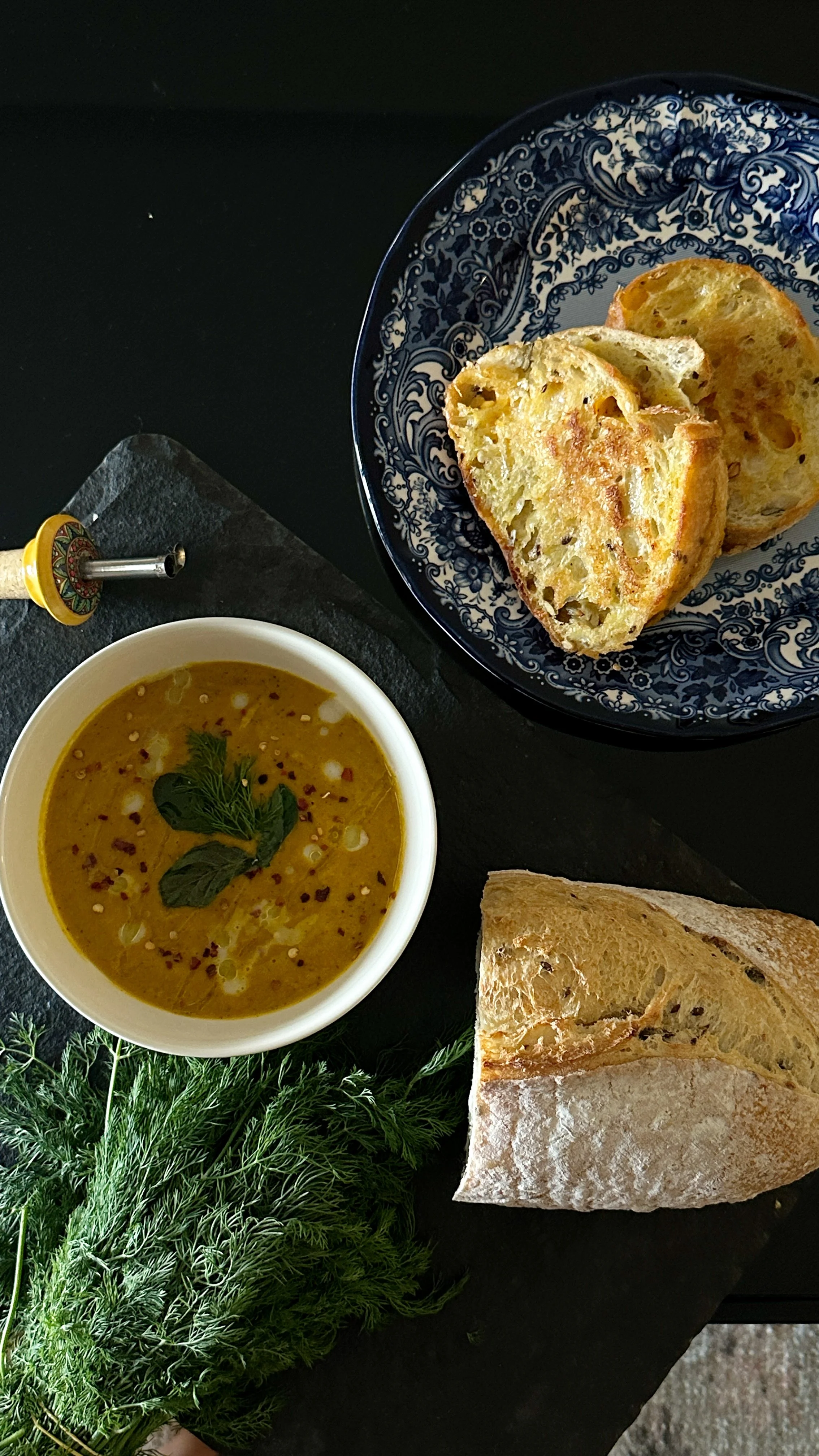 Bowl of yellow soup garnished with cilantro and spices, accompanied by slices of toasted bread and a crusty baguette on black surface with green herbs.