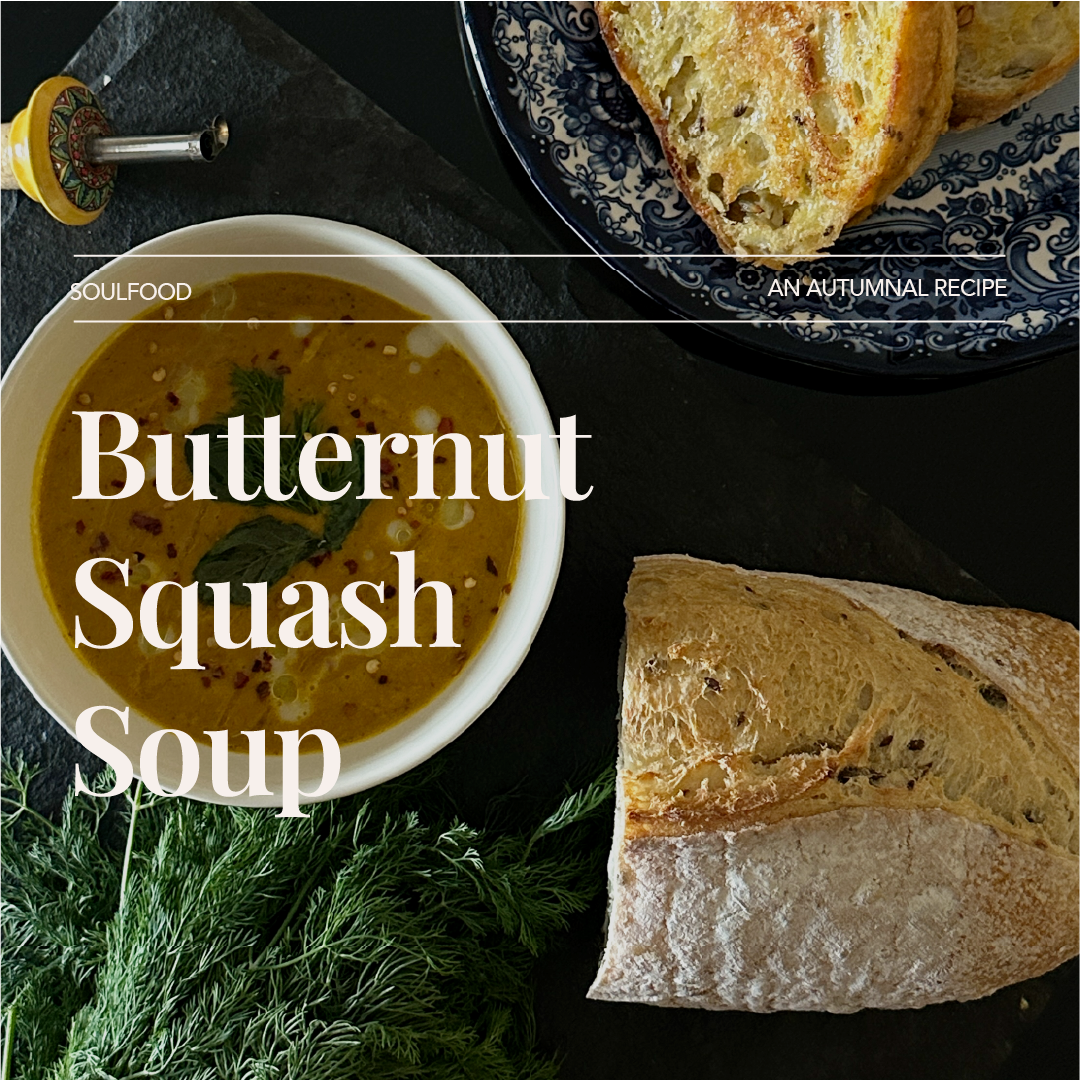 A bowl of butternut squash soup garnished with herbs, accompanied by slices of bread on a dark surface with fresh herbs and a decorative blue patterned plate in the background.
