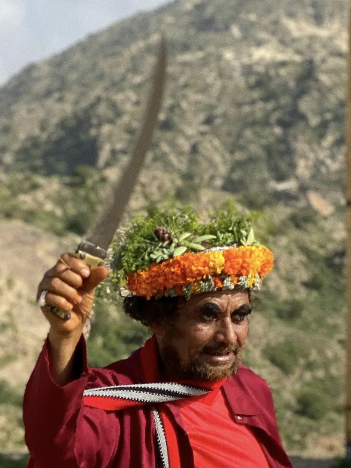 A man holding a large knife, wearing a flower crown, in a mountainous outdoor setting.