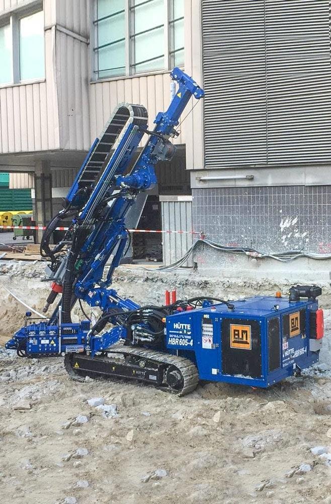 Small blue excavator with a long articulated arm at a construction site, surrounded by dirt and gravel, near a building with horizontal metal siding and windows.