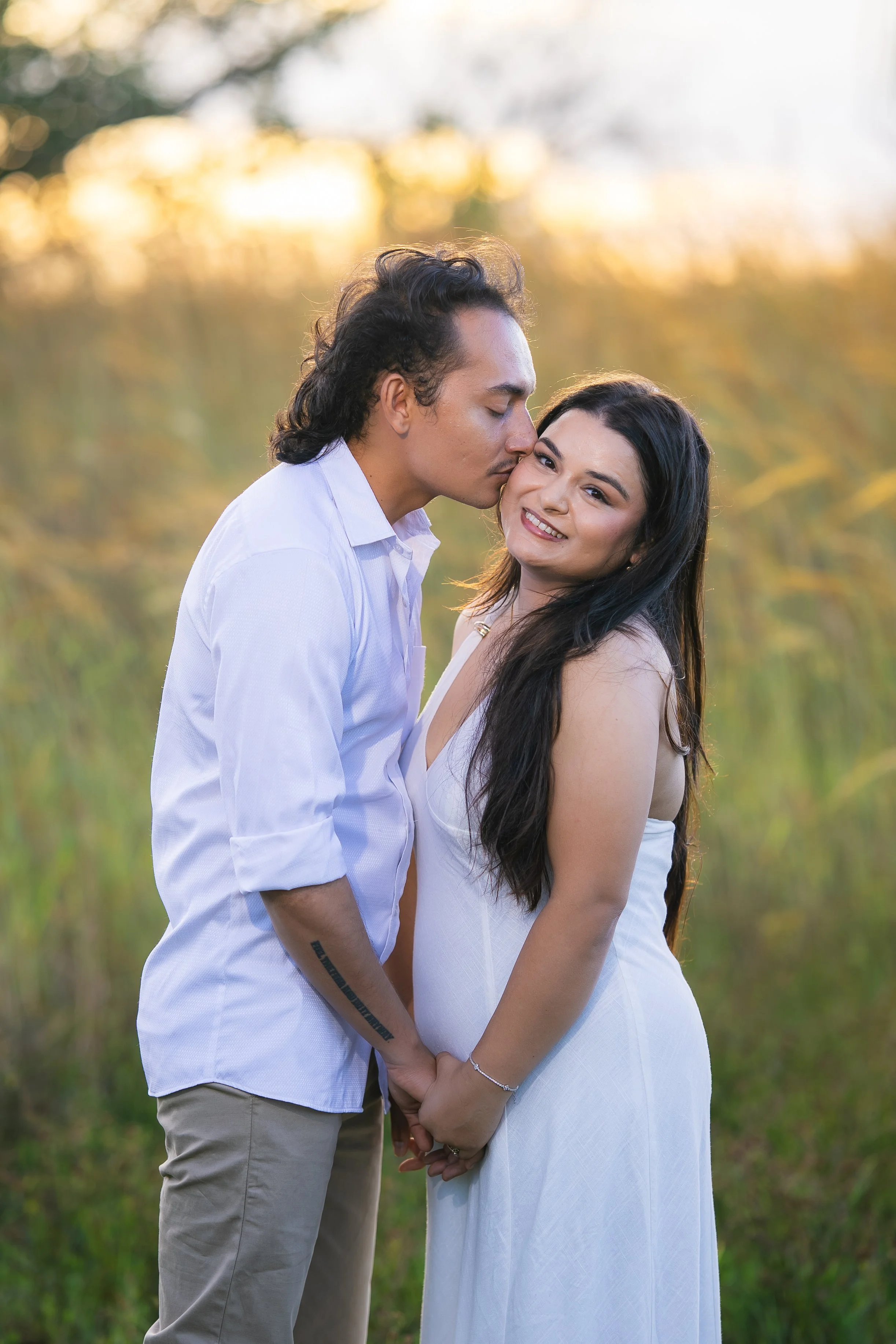 A couple is standing outdoors, holding hands, with the man kissing the woman's forehead. The woman is smiling warmly at the camera, both dressed in light-colored clothing, with a blurred natural background and warm sunlight.