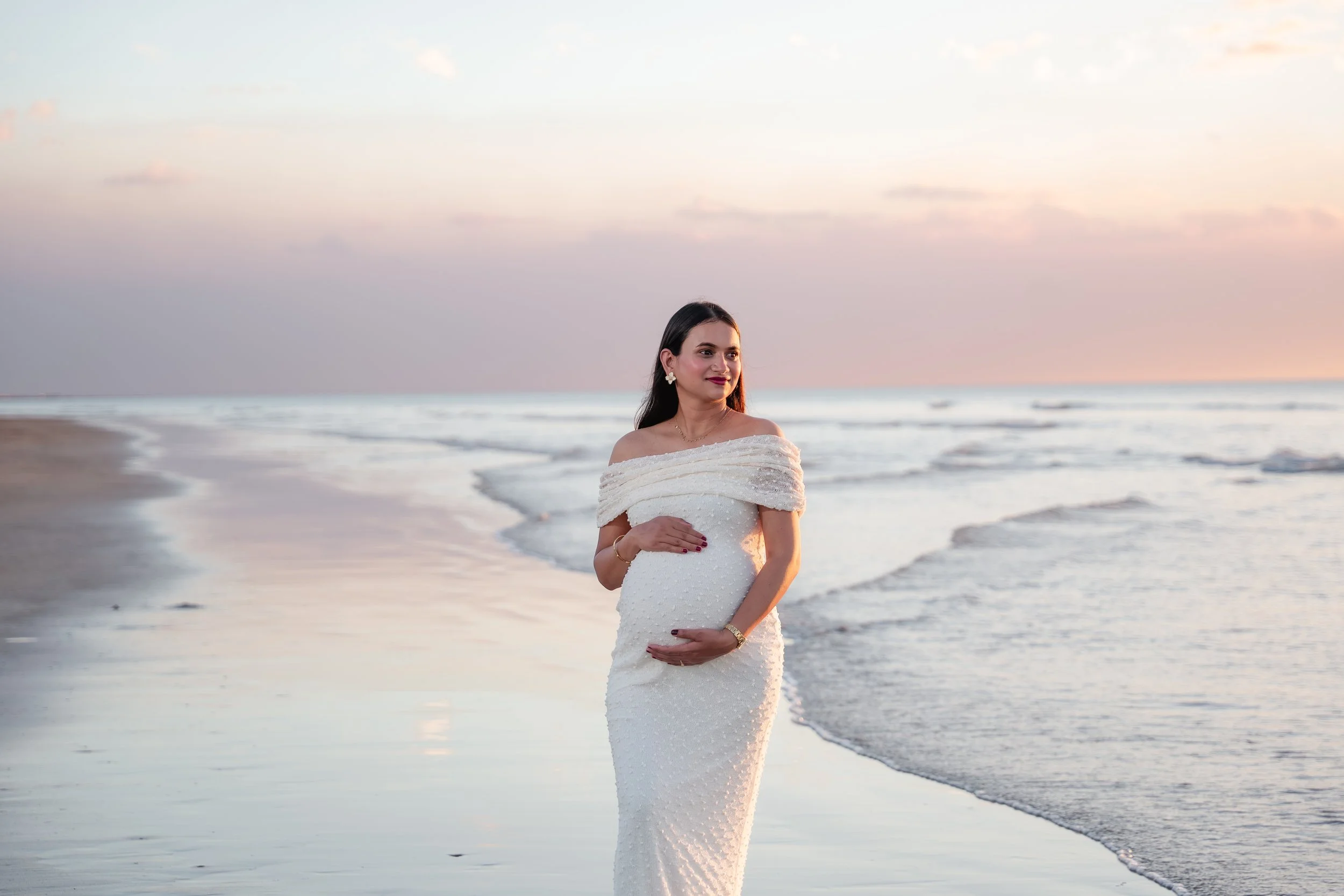 A woman in a white wedding dress standing on a beach at sunset, with the ocean and a colorful sky in the background.