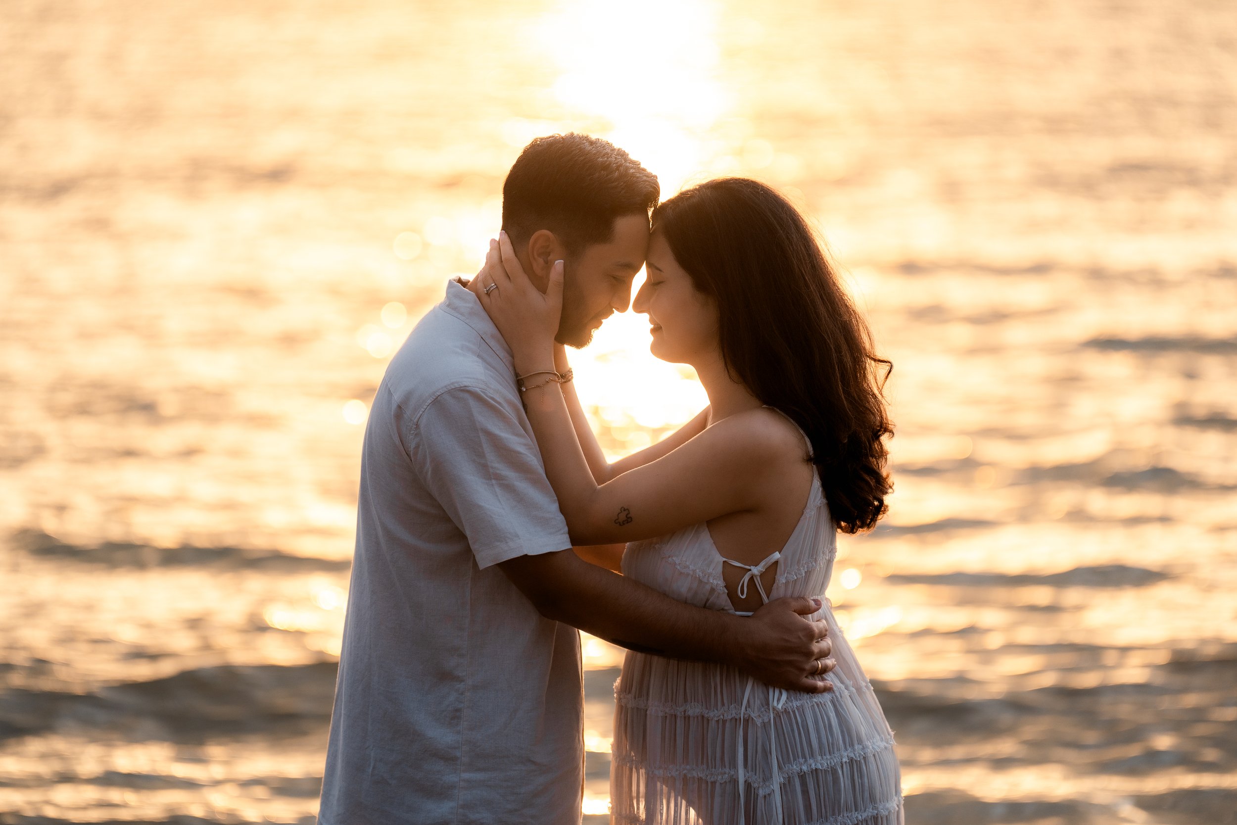 A man and woman embracing by the water at sunset, with heads touching and eyes closed, creating a romantic scene.