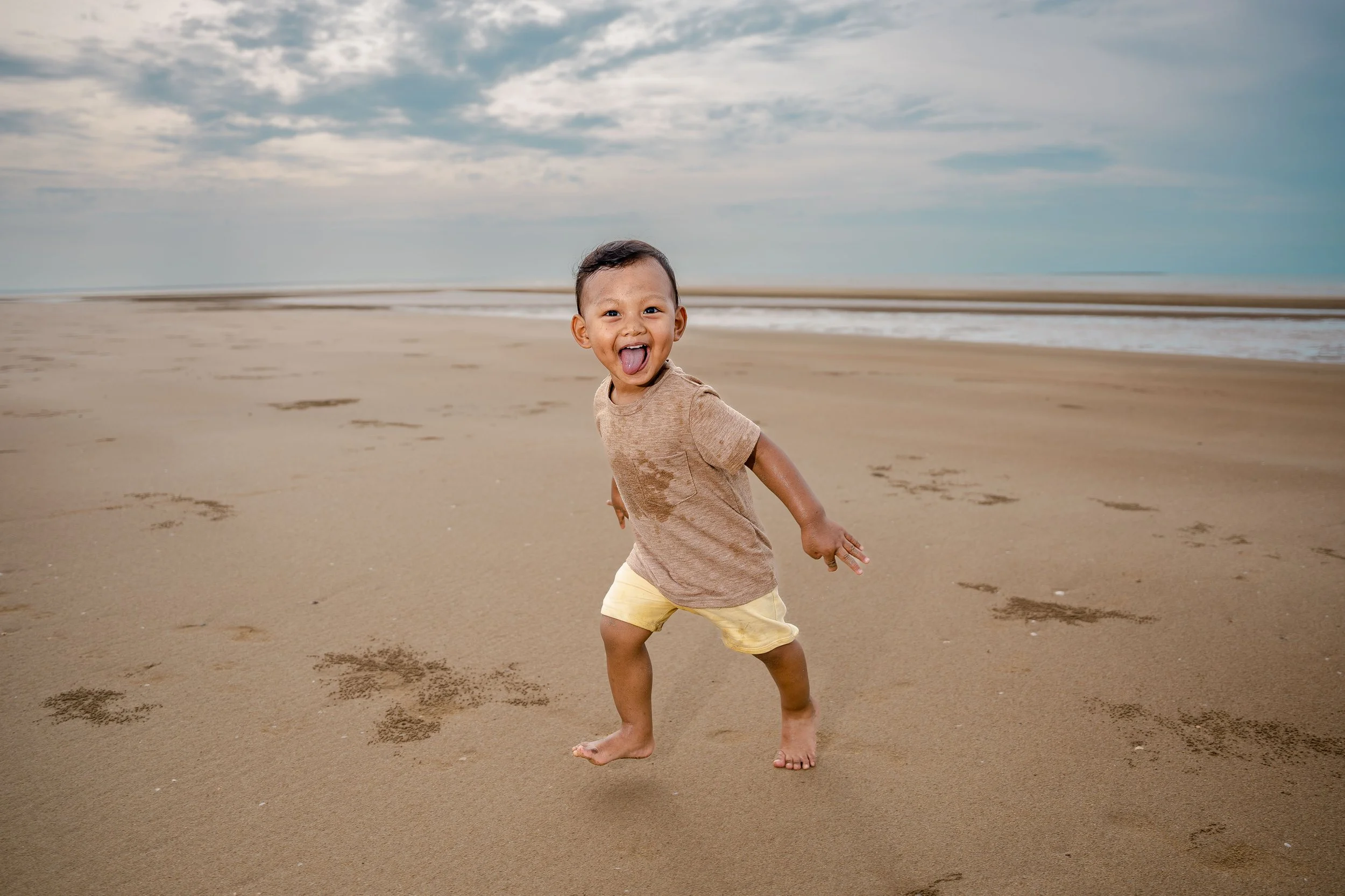 A young boy running barefoot on a sandy beach, smiling and sticking out his tongue, with clouds in the sky and the ocean in the background.