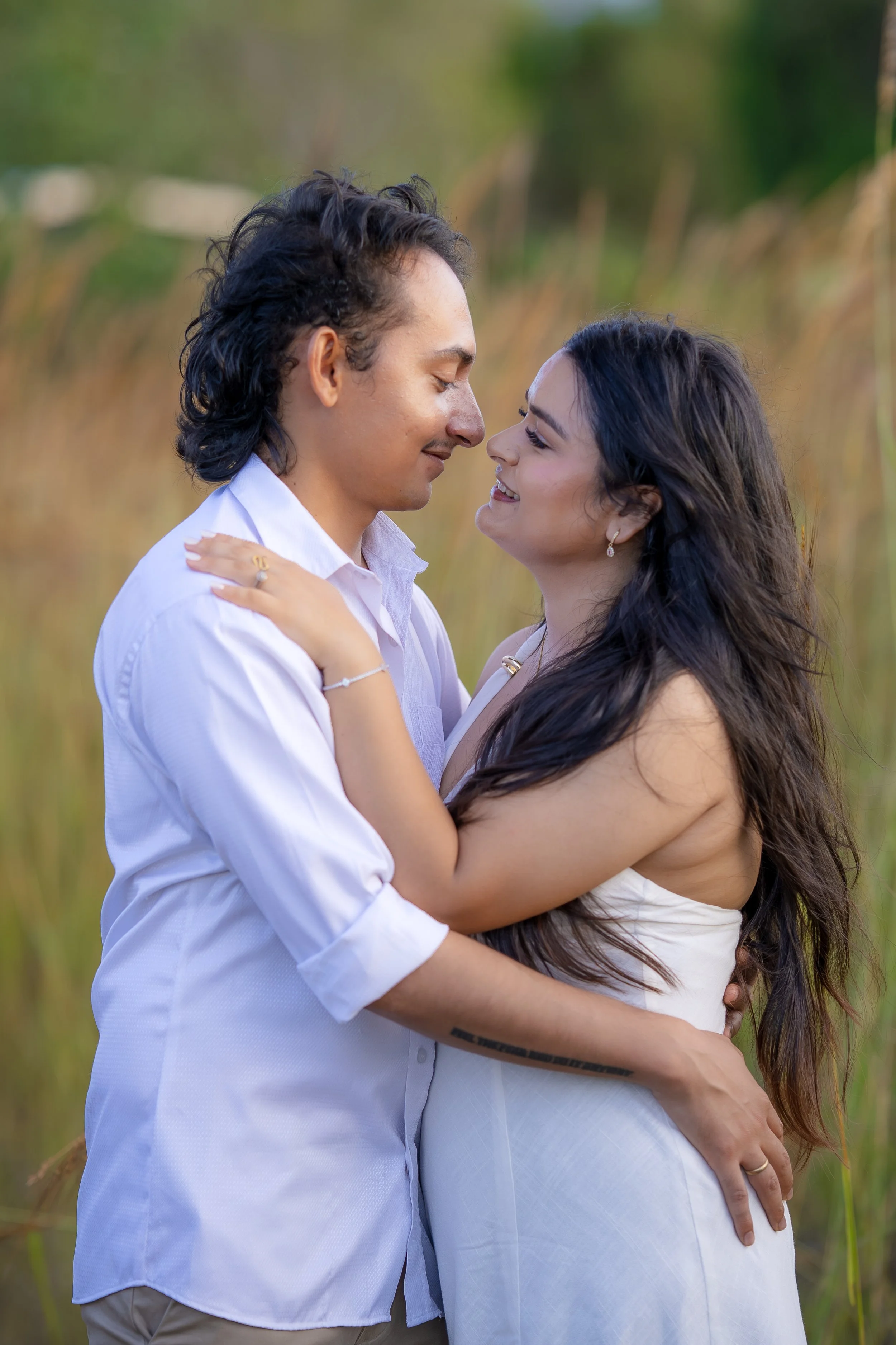 A man and a woman are embracing outdoors in a grassy field, facing each other closely with smiles, with the man gently holding the woman around her shoulders and the woman with her arms around the man's neck.