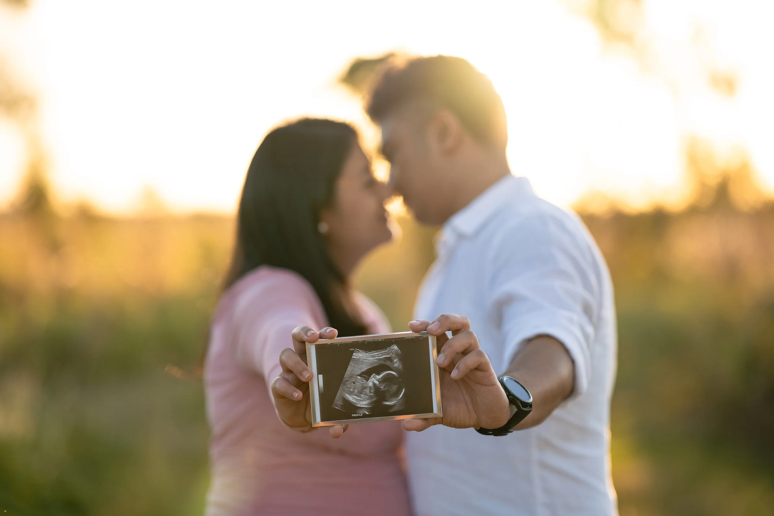 A couple stands close together outdoors during sunset, holding an ultrasound picture while a woman prepares to take a selfie with her partner.