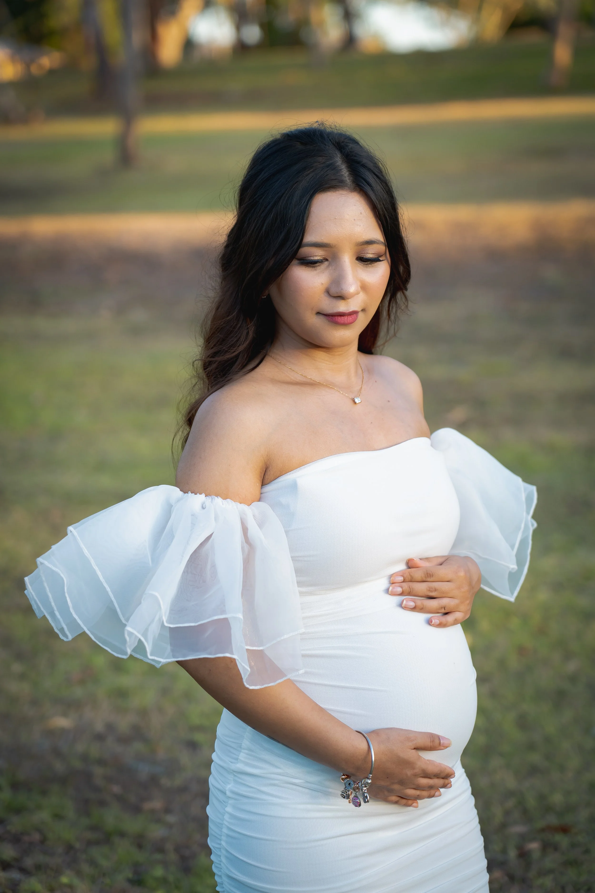 A pregnant woman with dark hair and tan skin wearing a white off-shoulder dress and jewelry, standing outdoors in a park-like setting during sunset.