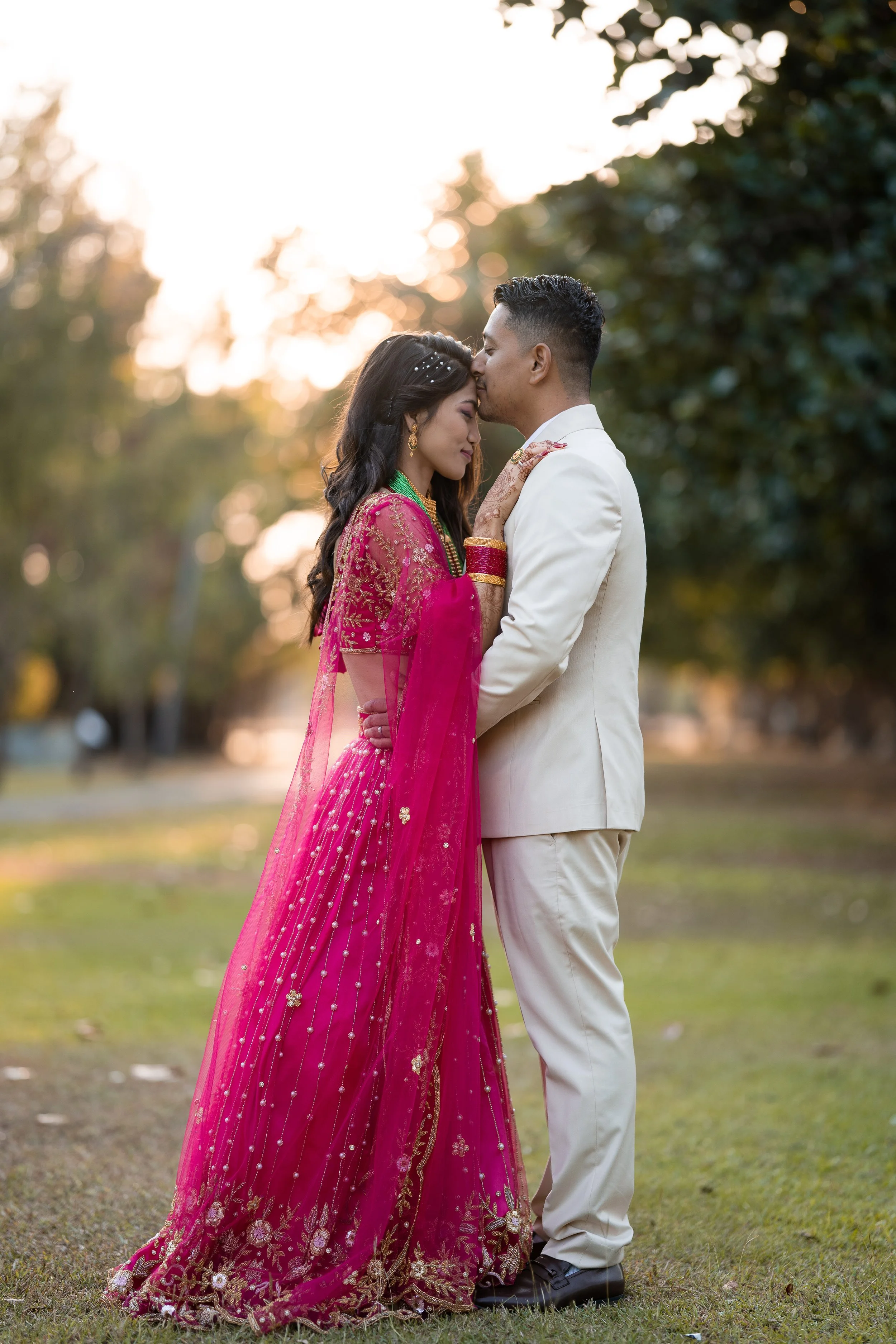A couple in formal attire, the woman wearing a pink embroidered saree and the man in a white suit, stand closely together outdoors during sunset, with trees in the background.
