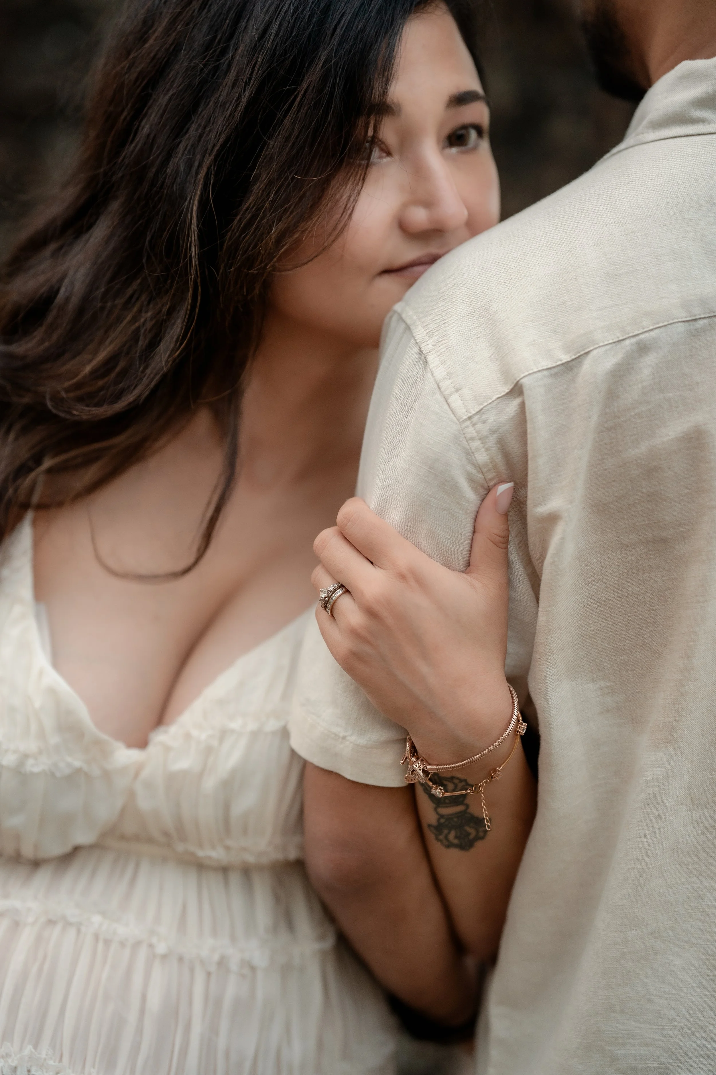 A woman with long, wavy dark hair, wearing a white dress, embraces a man in a light-colored shirt. She has her hand on his shoulder, showcasing rings and bracelets. She is looking back with a gentle expression.