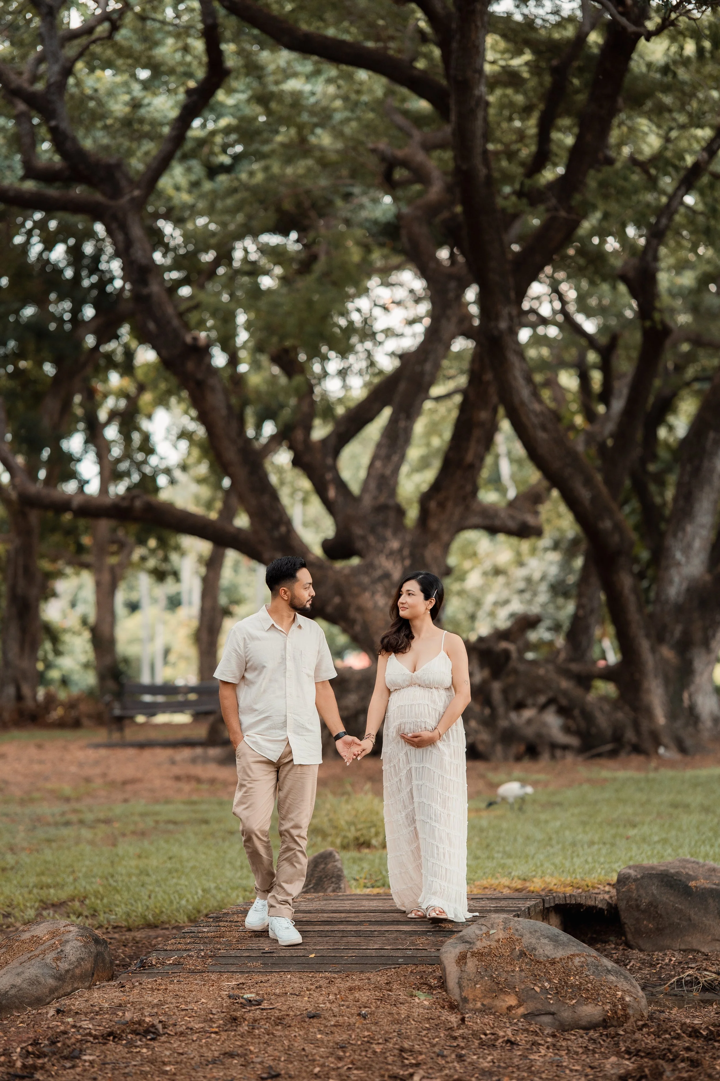 A couple walks hand in hand on a wooden bridge in a park with large trees and green grass. The woman is pregnant and wears a white dress. The man wears a light shirt and beige pants. They look at each other affectionately.