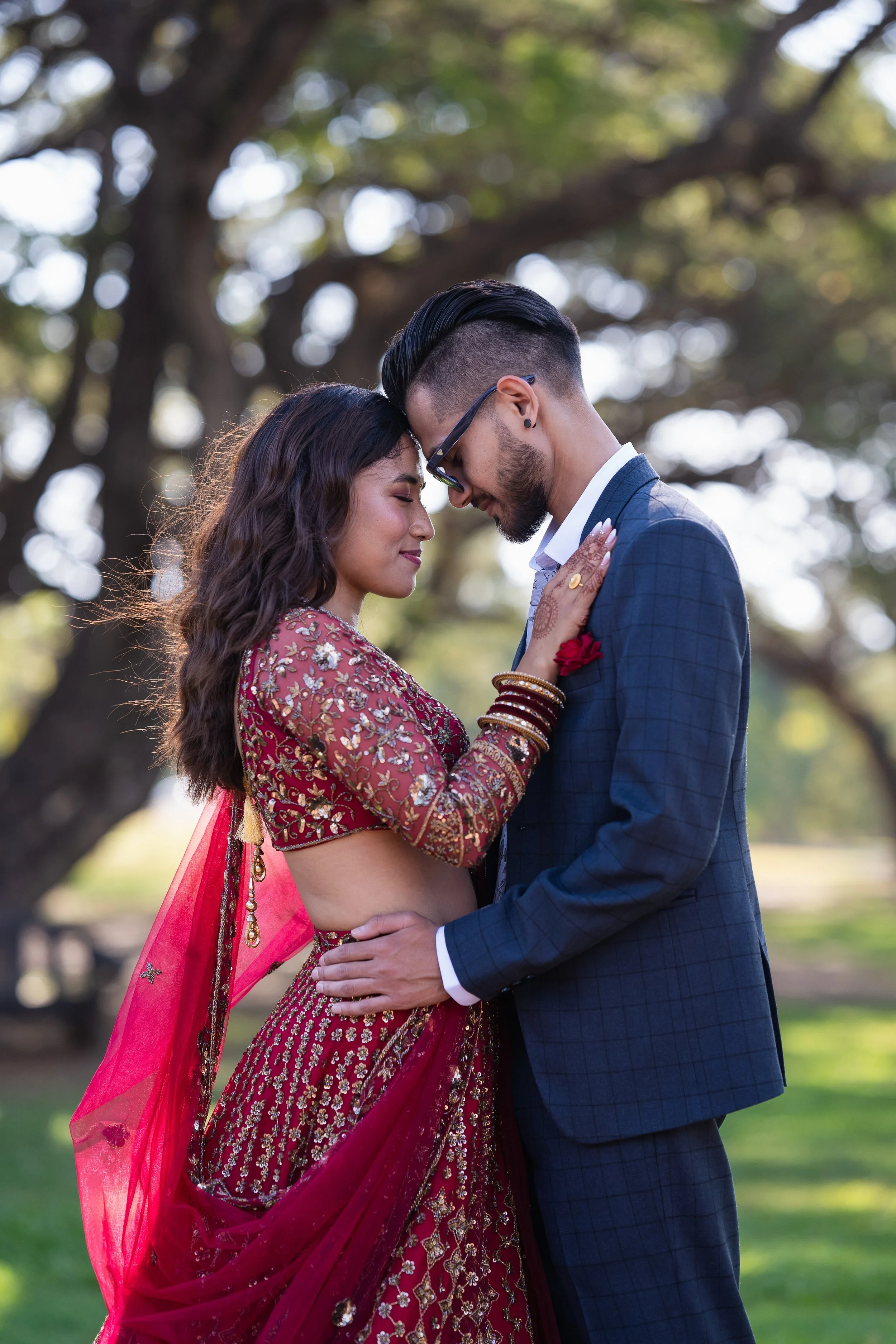 A couple is standing close together outdoors, with their foreheads touching and eyes closed. The woman is wearing a colorful, embroidered traditional dress and headscarf, while the man is dressed in a blue suit with a white shirt. There are large tre