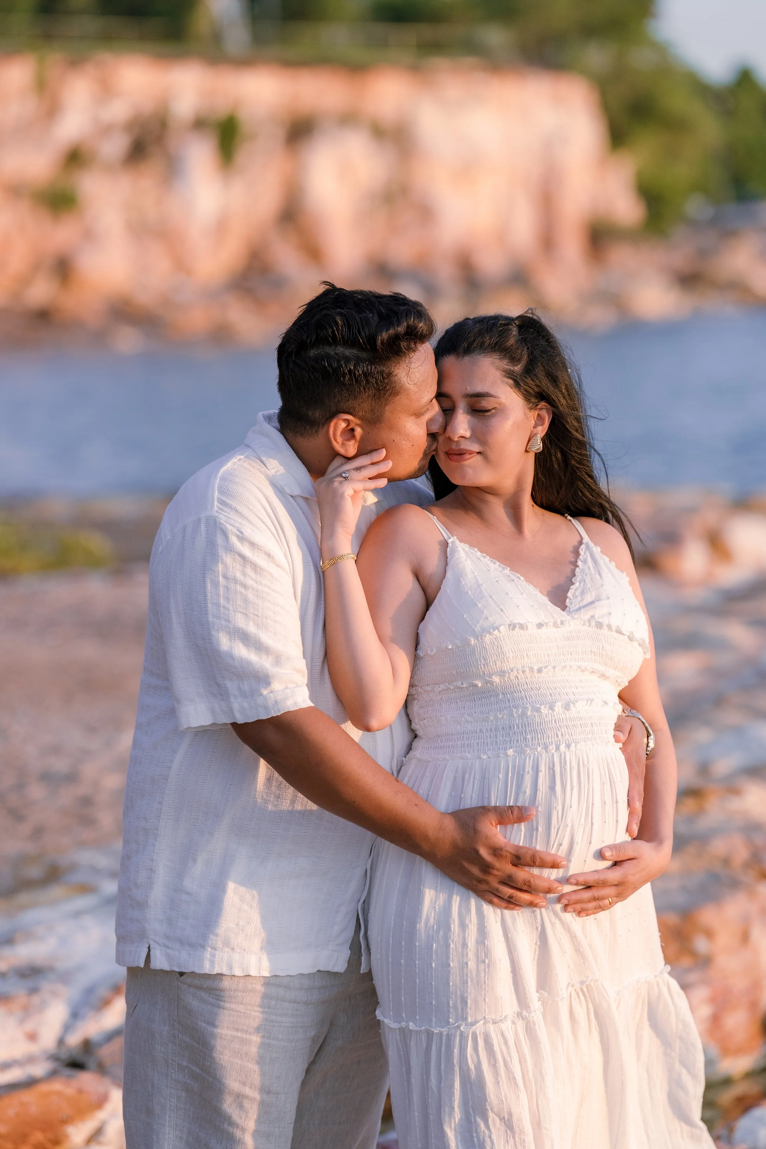 A couple stands on the beach, with the man touching the pregnant woman's belly, and they are about to kiss. The woman is wearing a white dress and the man is wearing a white shirt. There is a rocky cliff and water in the background.