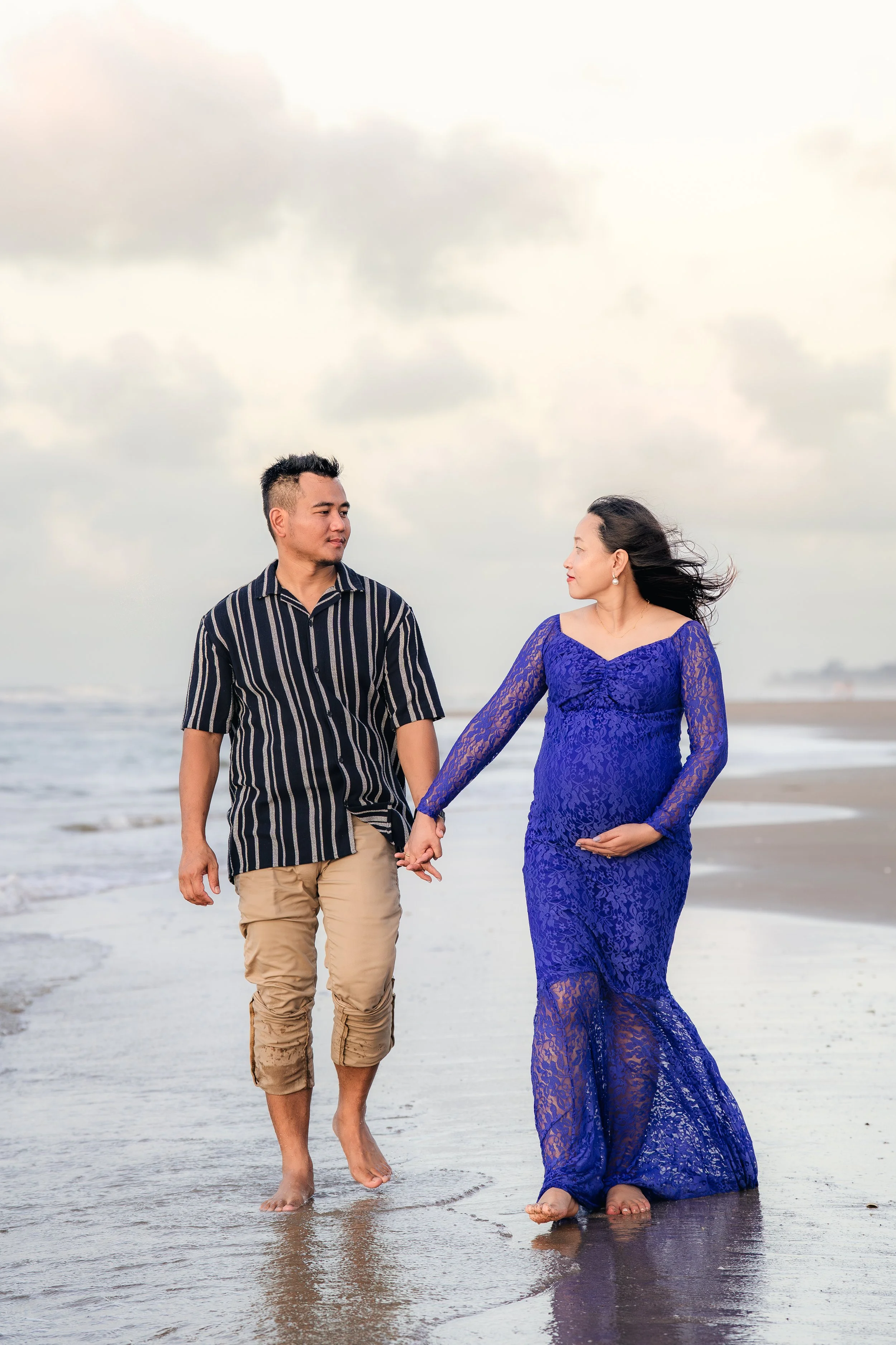 A man and woman holding hands and walking barefoot on the beach with cloudy sky overhead. The woman is wearing a royal blue lace dress and the man is wearing a black and white striped shirt with khaki pants.