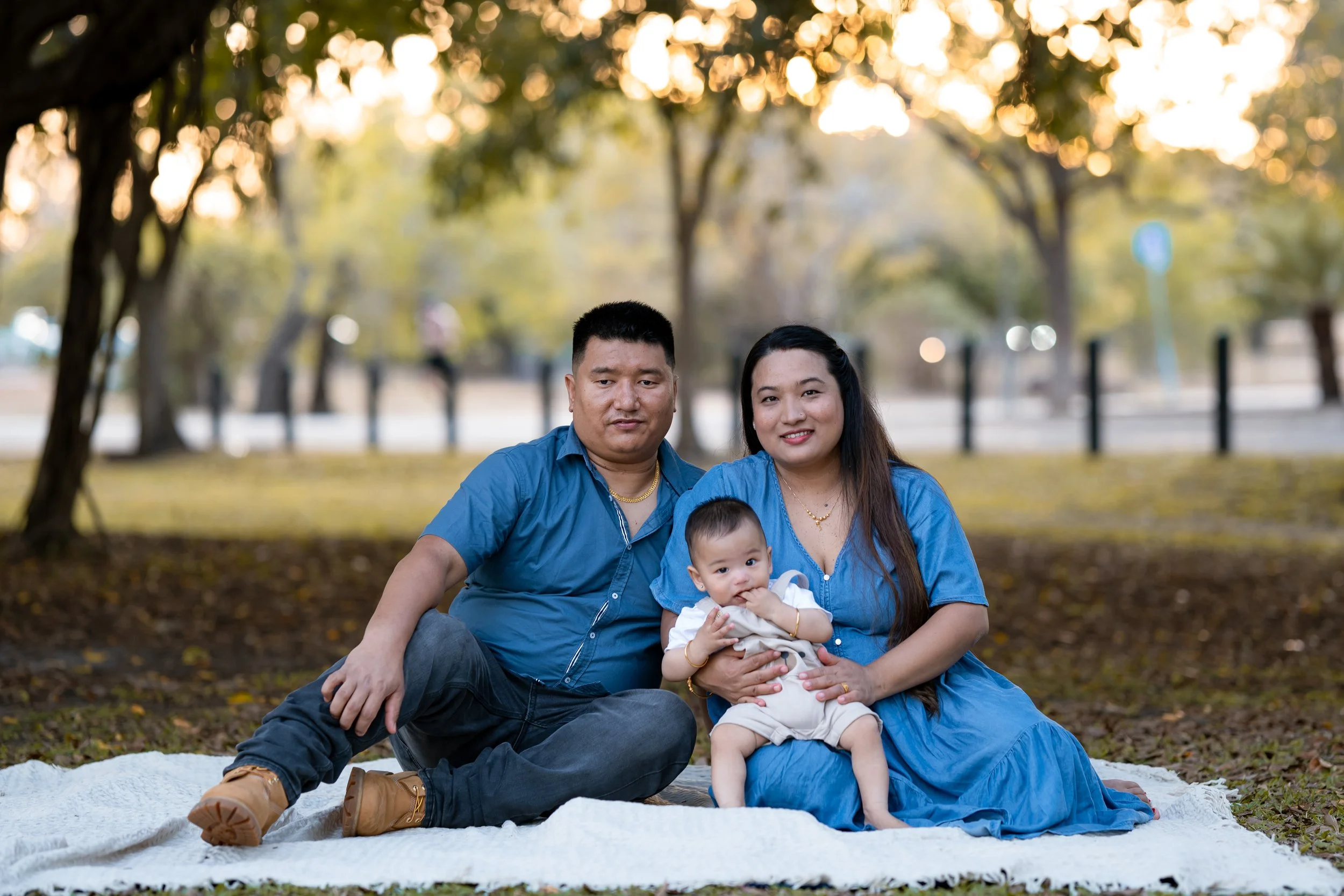 A family of three sitting outdoors on a blanked in a park during fall, with trees and autumn leaves in the background. The man, woman, and baby are all smiling at the camera.