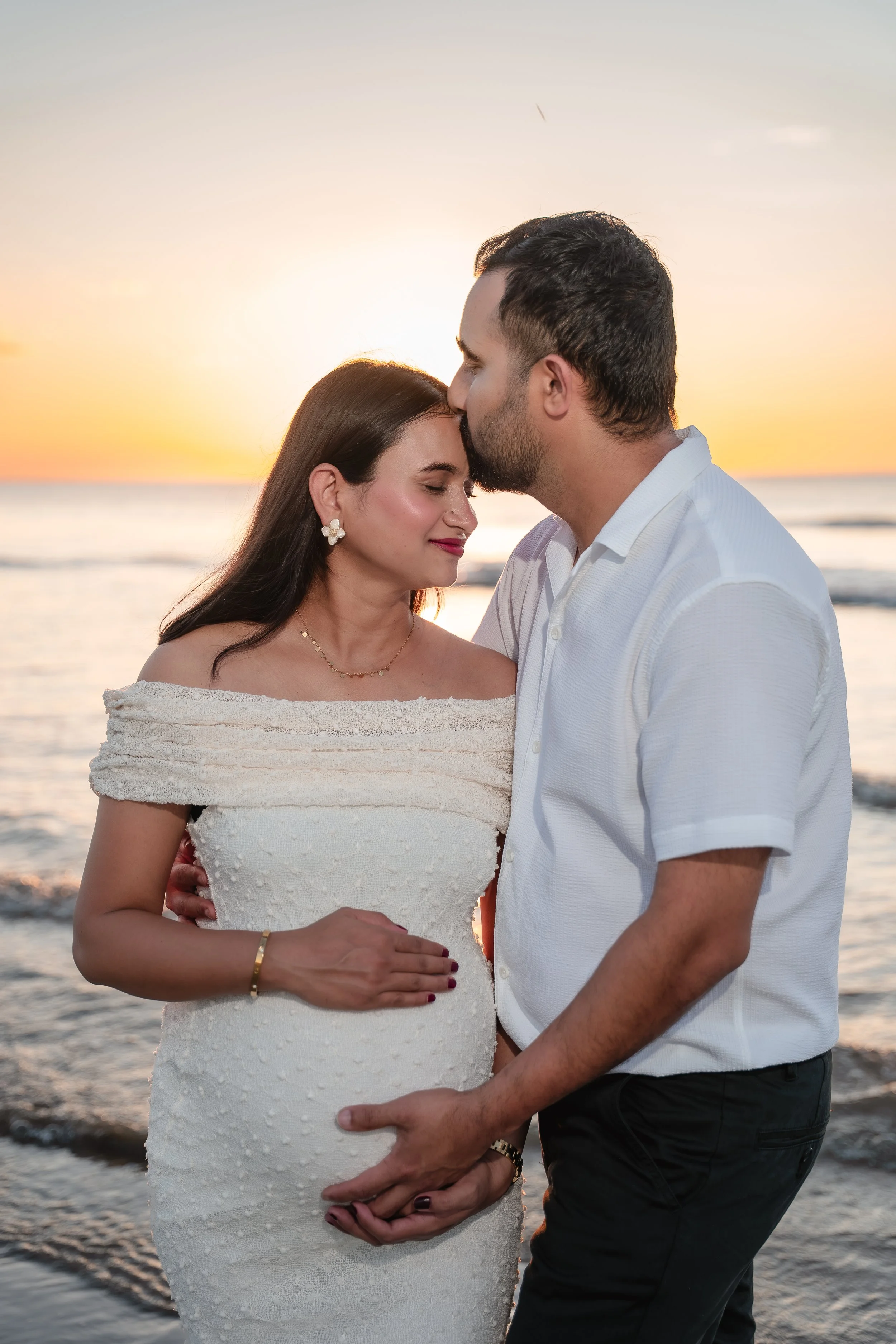 A couple stands on the beach at sunset, with the man kissing the woman on the forehead and both holding each other's hands, the woman smiling gently with eyes closed.