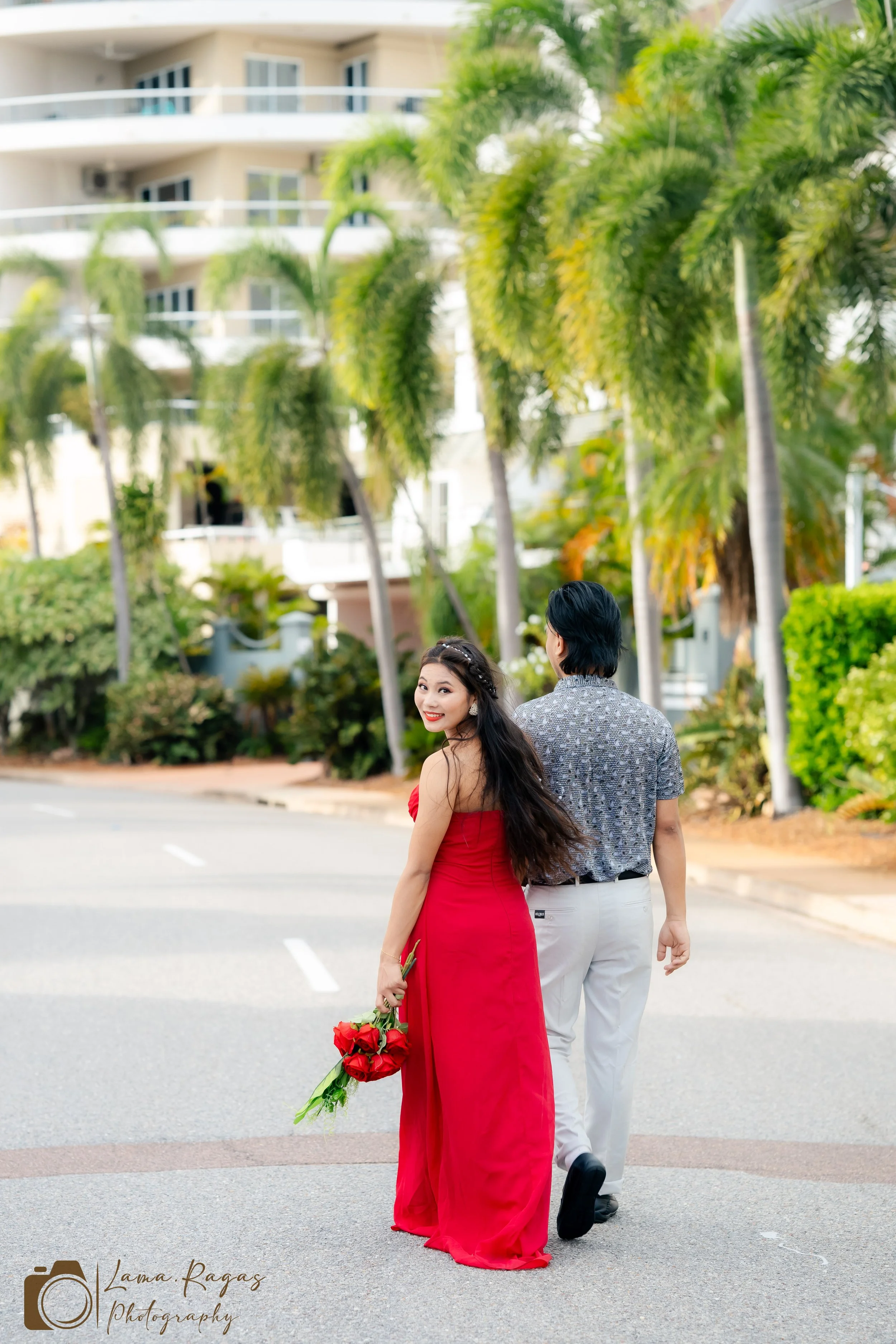 A couple walking on a street, the woman wearing a red strapless gown holding a bouquet of red roses, the man in a grey patterned shirt and white pants, with a background of palm trees and a beige building with balconies, during daytime.