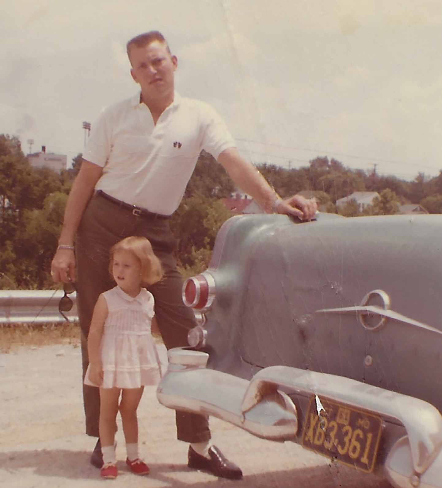 Picture of a man standing with his hand on the trunk of a car from the 1960s and a little girl standing in front of him with her arm around his leg. They are standing on the side of a road with a city landscape behind them.