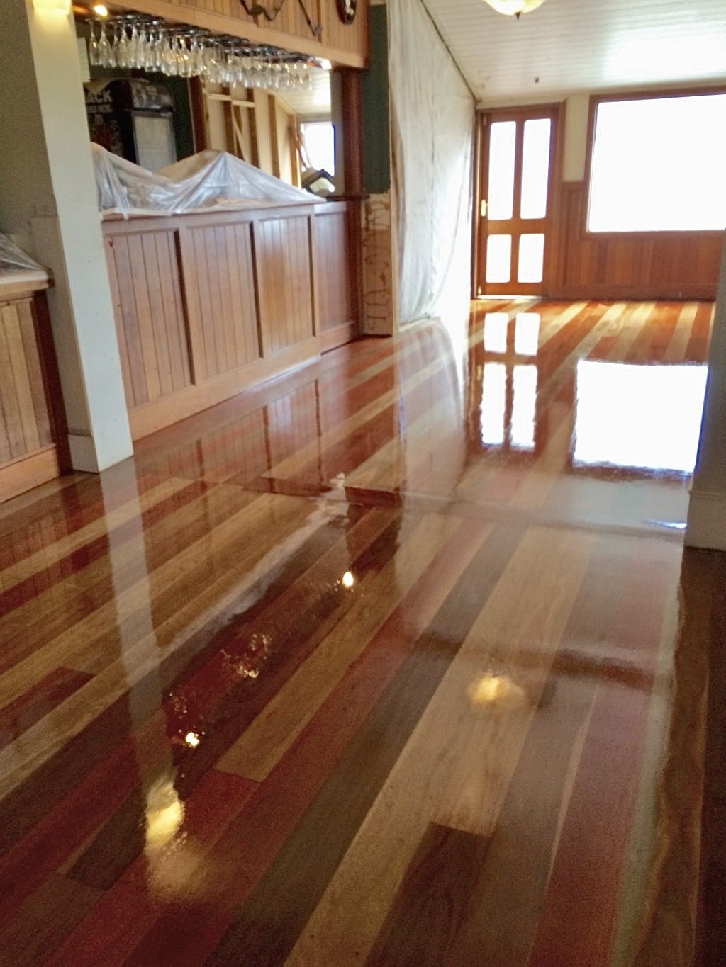 Interior of a room with shiny wooden floors, wooden cabinets, glass hanging above the bar area, large windows, and a door leading outside.