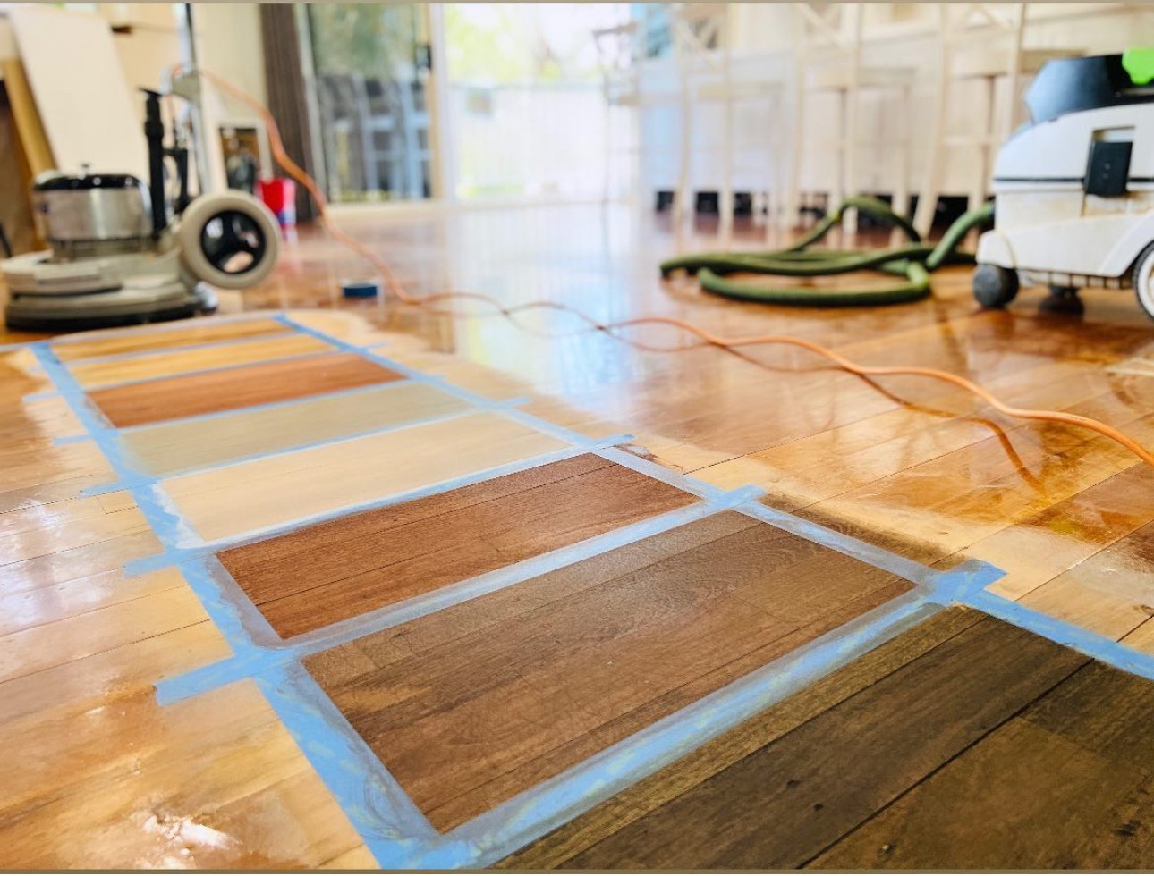 Flooring samples in various shades of wood stain laid out on wooden floor, with sanding and finishing tools, and construction equipment nearby.