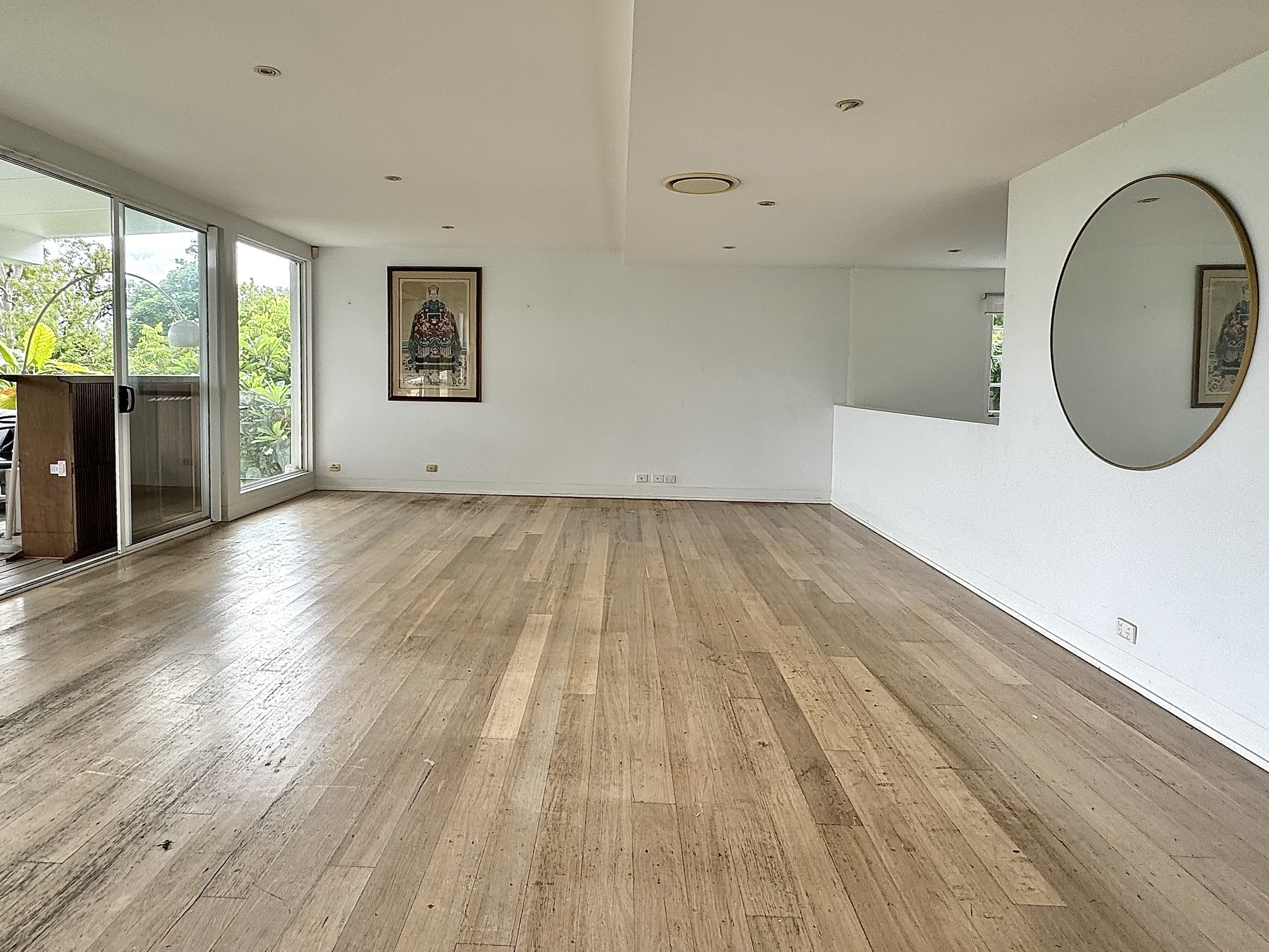 An empty living room with light wood floors, white walls, a large round mirror on the right wall, a framed picture on the far wall, and a sliding glass door leading to a balcony with greenery outside.