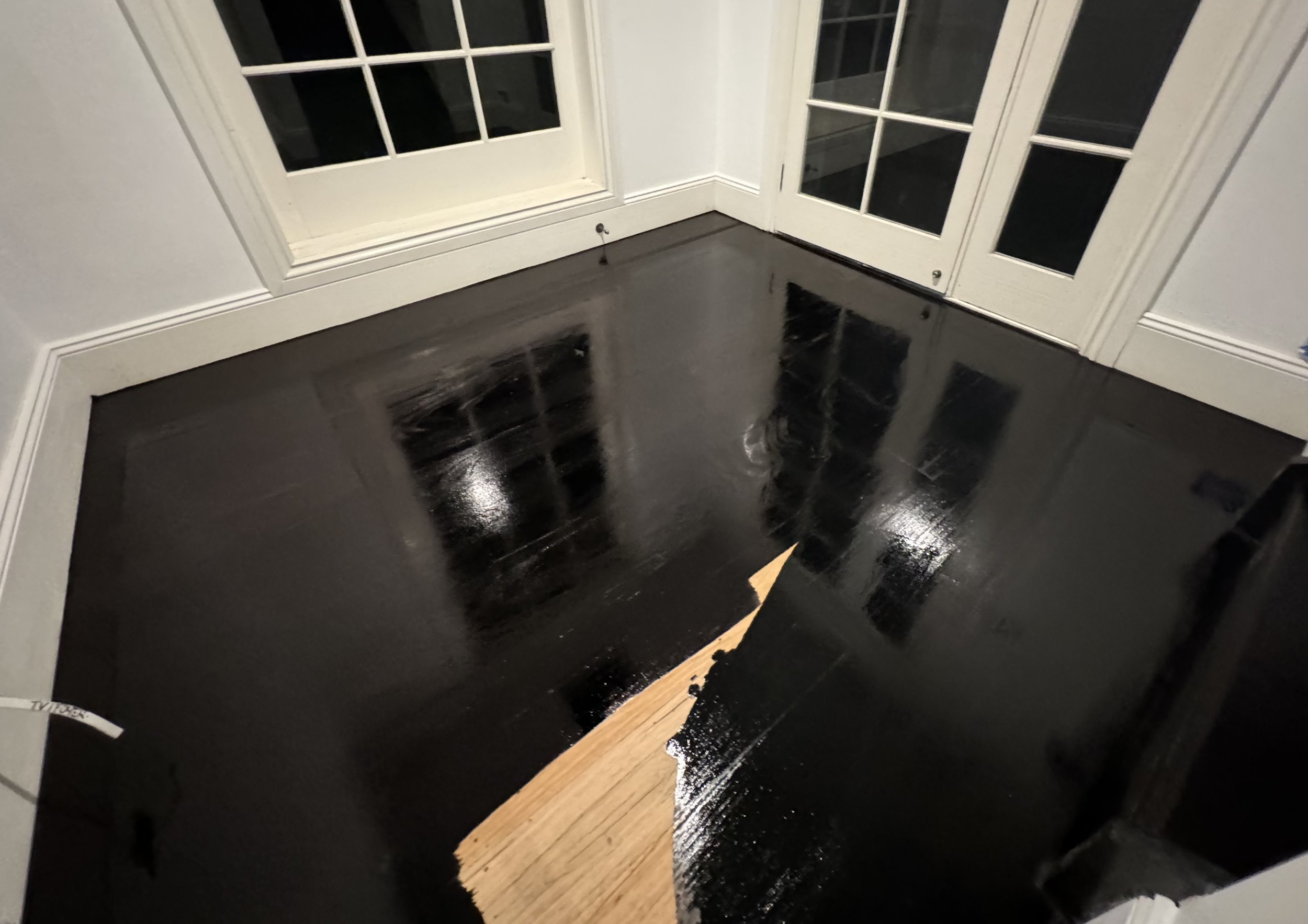 Room with black glossy painted floor, a small section of unfinished wood flooring, and white walls with French doors.