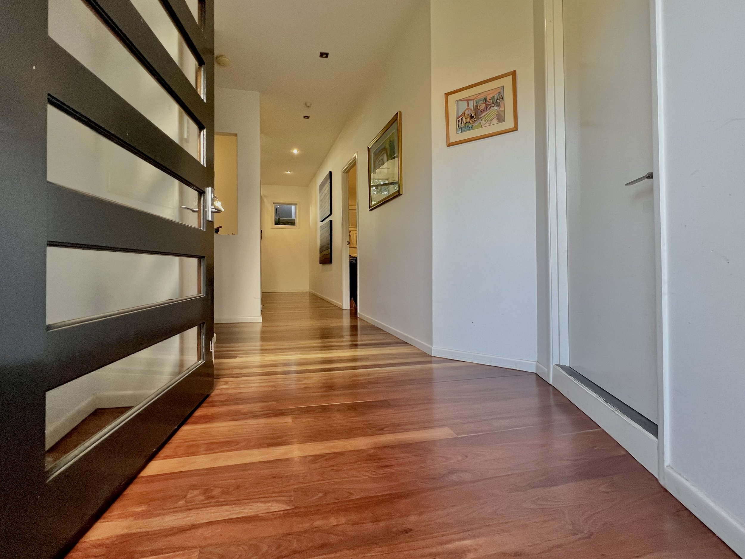 View of a hallway with hardwood floors, white walls, and framed artwork, taken from a low angle near a black railing or partition on the left side, leading towards a door at the end.