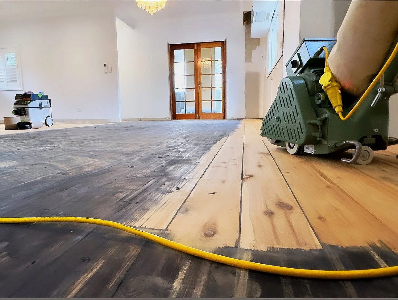 Room undergoing hardwood floor refinishing with a sander, showing partly sanded and unsanded wood floors, with equipment and yellow extension cord.