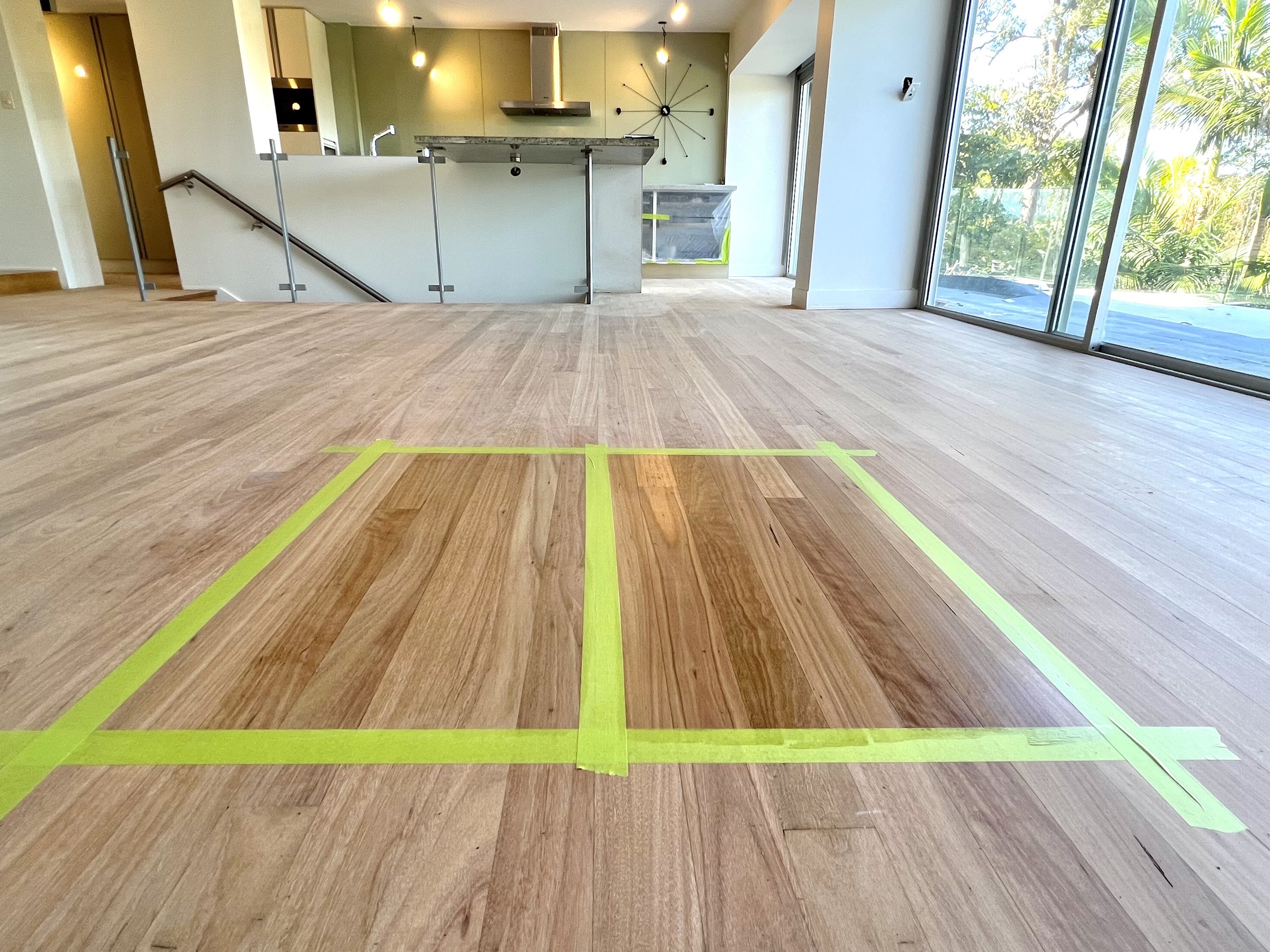 Empty room with wooden flooring, marked with yellow tape for measurement or installation, and a kitchen area with an island, large glass sliding doors, and a balcony outside.