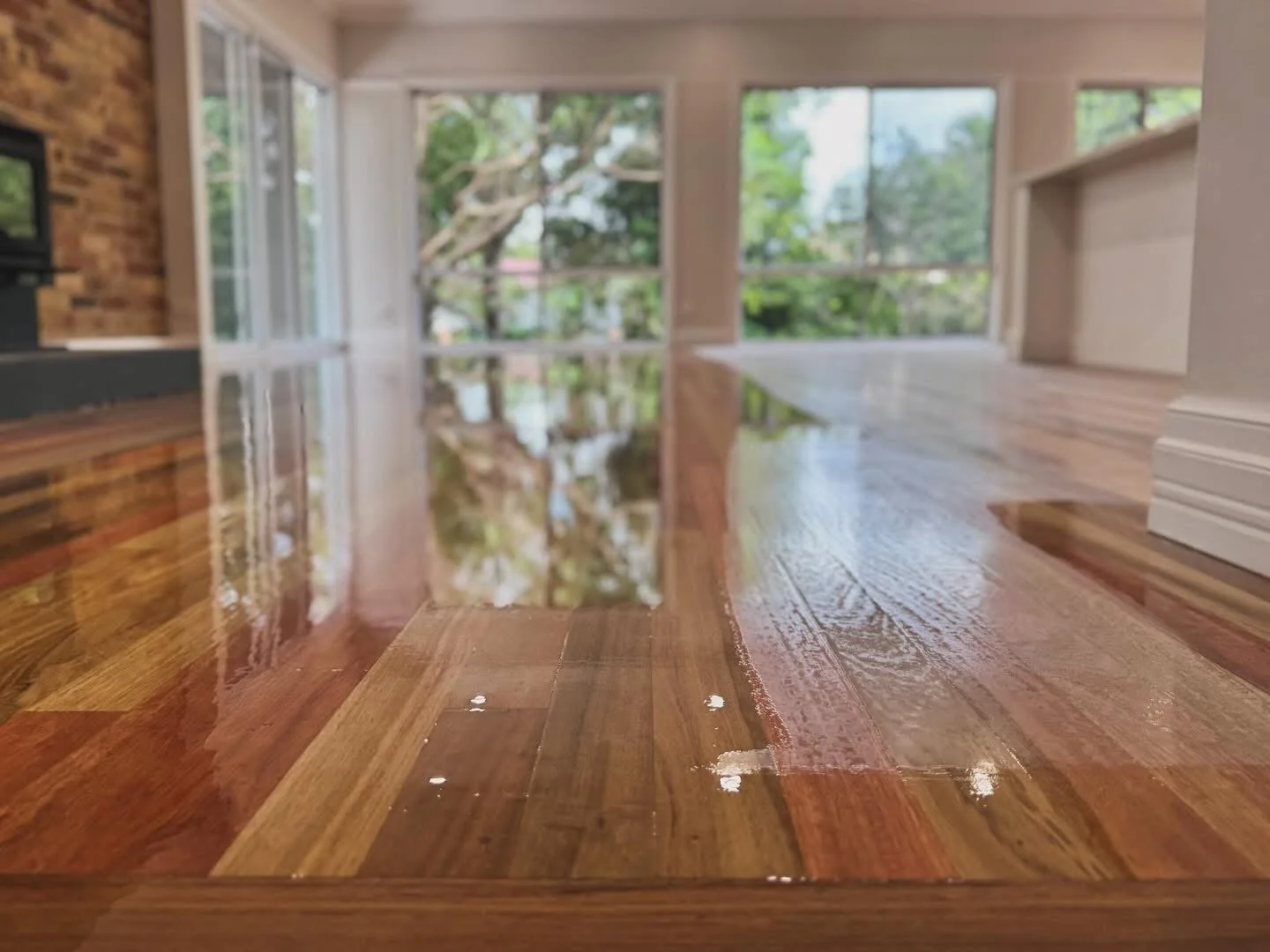 Timber floor polishing Brisbane view of a shiny wooden floor in a bright room with large windows showing green trees outside.