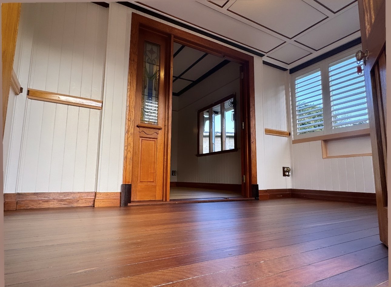Empty room with wooden floors and trim, white panels on the walls, and windows with white shutters letting in natural light.