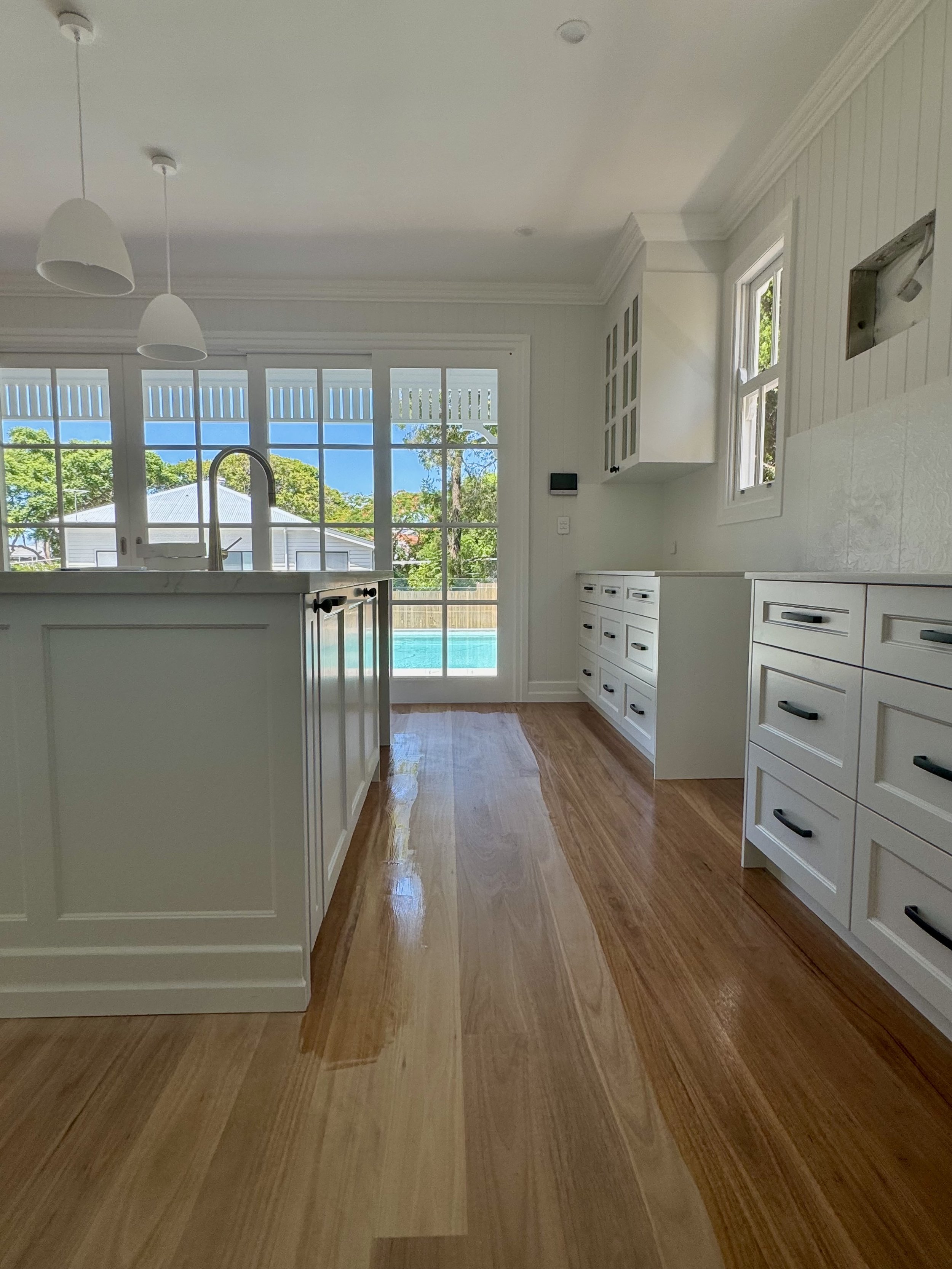Kitchen with white cabinets, wooden floor, and large windows showing a pool and backyard outside.