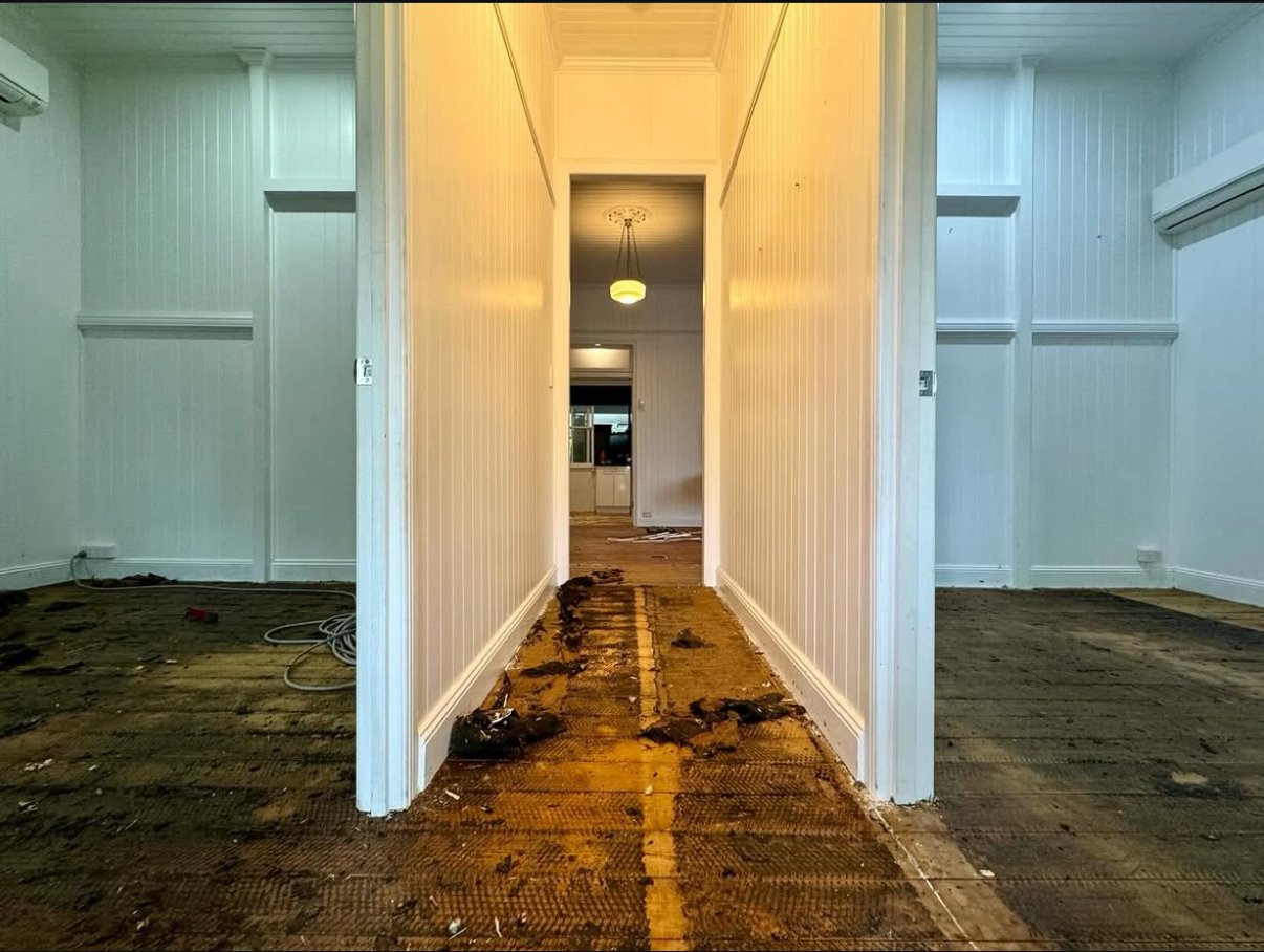 Interior of a house under renovation with a damaged hardwood floor, with white walls and ceiling, and visible wiring on the left floor area in Brisbane based.