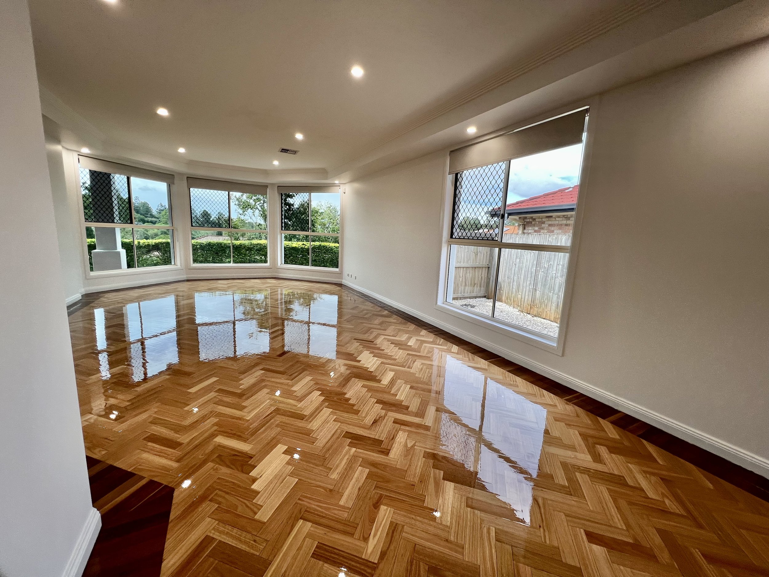 Empty living room with polished wooden parquet floors, large windows with security screens, and white walls.