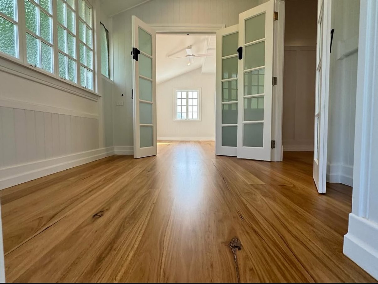 View of an empty room with wood flooring, white walls, and a set of French doors opening into a sunlit adjacent room with a window and ceiling fan.