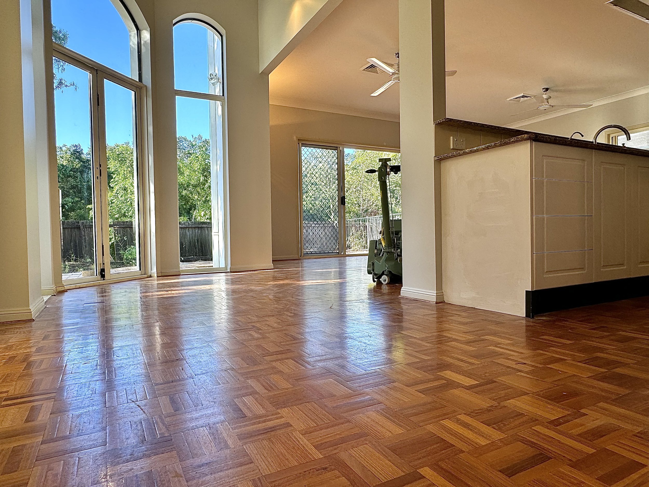 Empty living room with wood flooring, large windows, and sliding glass doors leading outside. Part of the kitchen area visible with a bar, ceiling fans, and sliding door with a screen door.