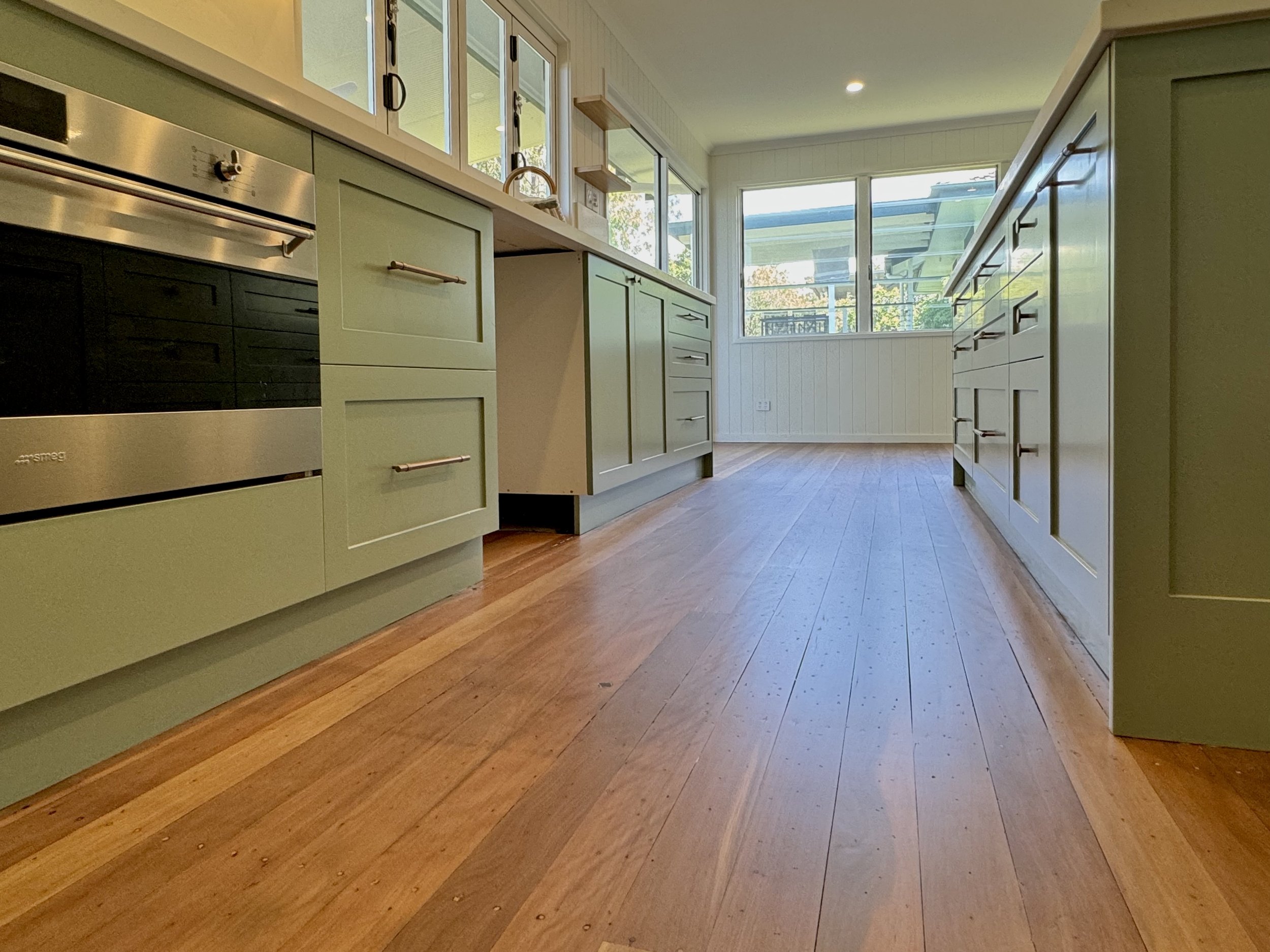 A bright, modern kitchen with light green cabinets, wooden flooring, and large windows providing natural light.