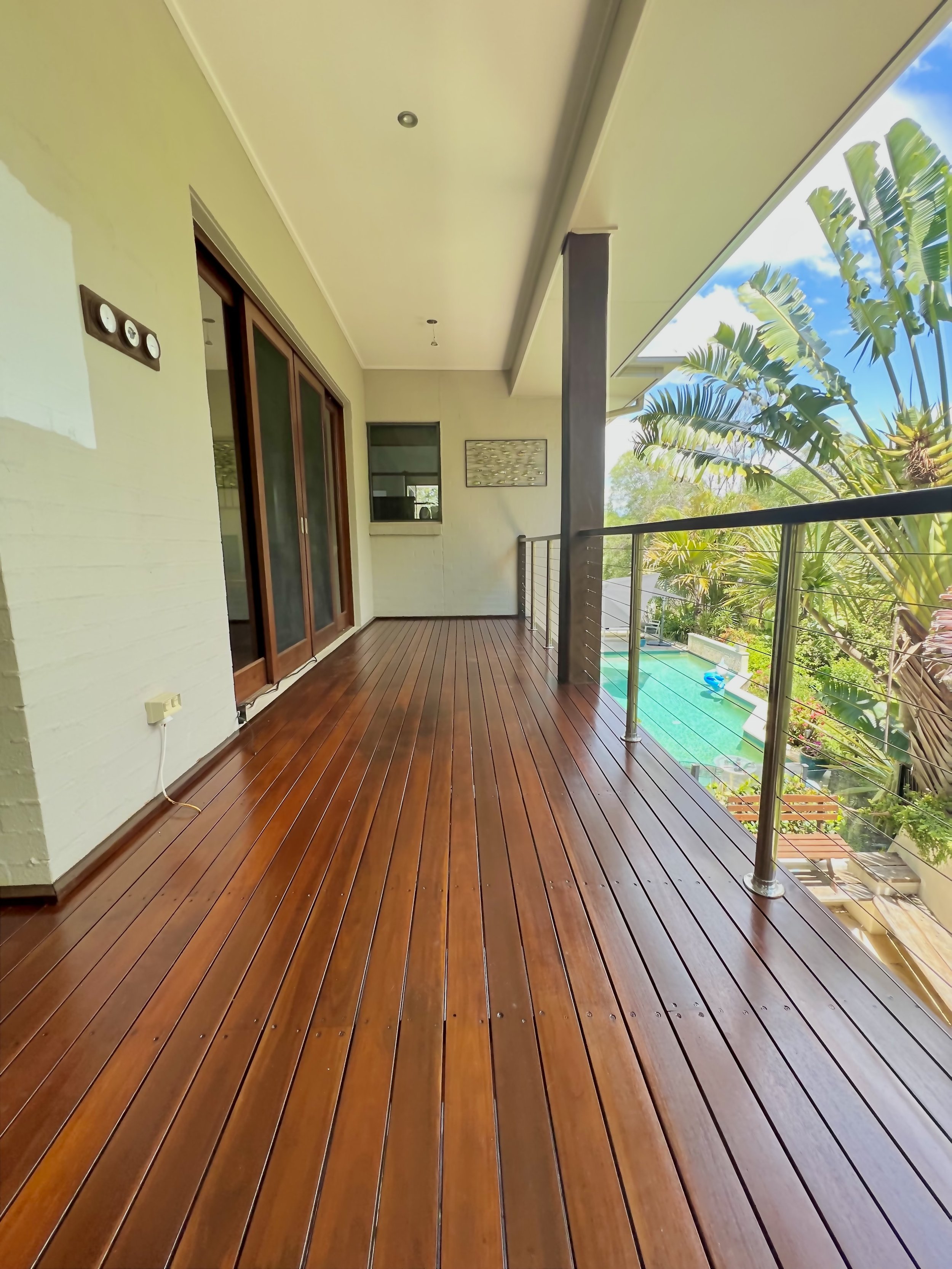Wooden balcony with a metal railing, overlooking a lush green garden with tropical plants and a swimming pool.