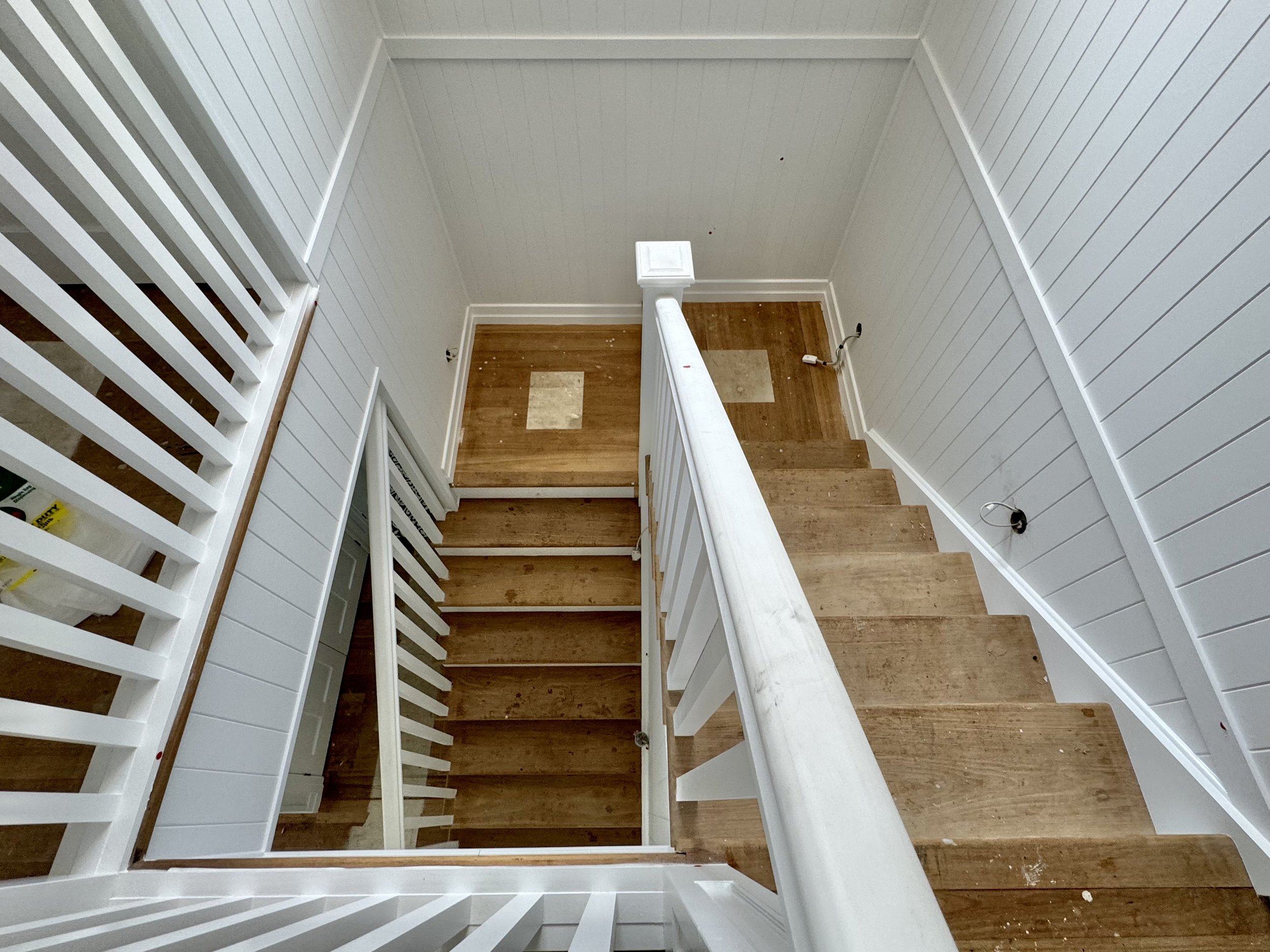 Interior view of a staircase with wooden steps, white handrail, and white panel walls, leading down to a small landing with a wooden floor. Electrical wires and outlets are visible on the walls.