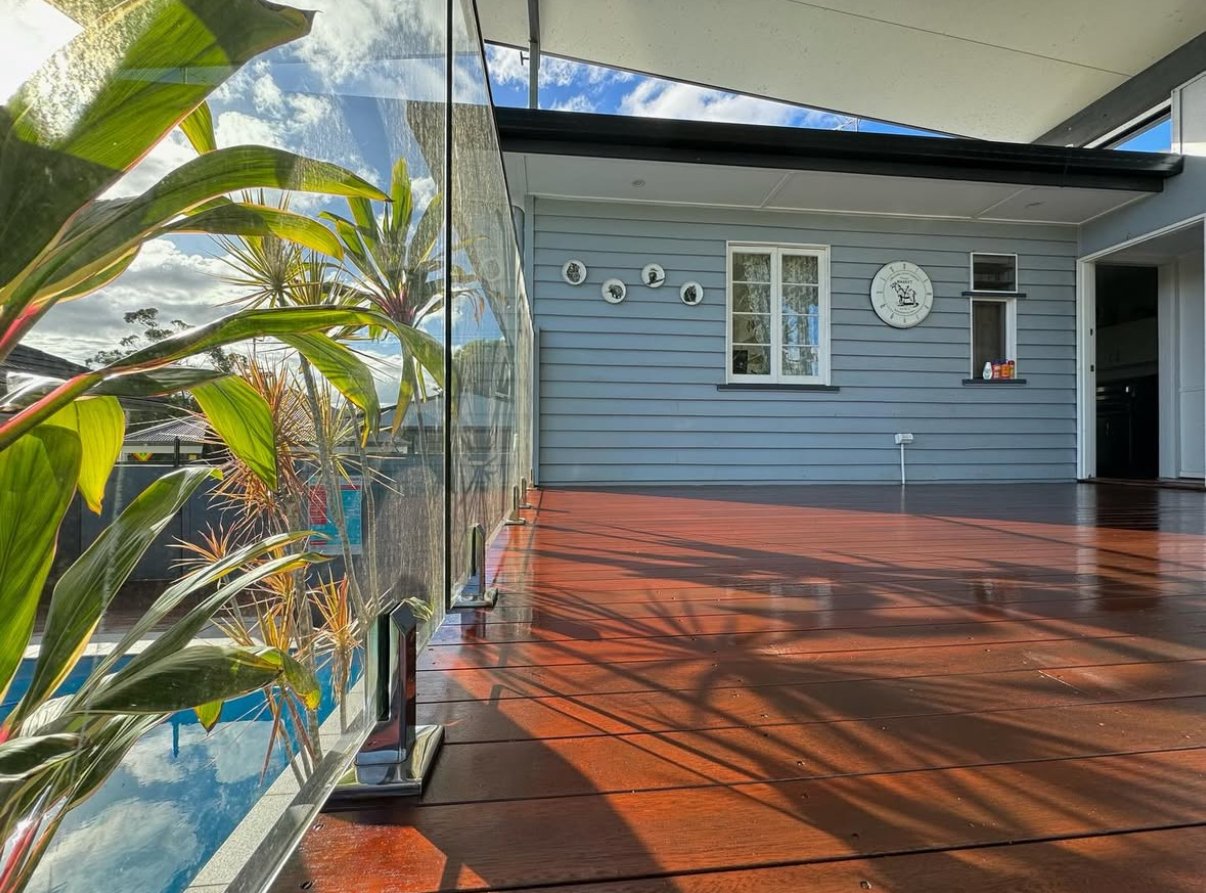 A wooden outdoor deck with a glass railing, reflected clouds and trees, and a blue house in the background. The house has decorative plates and a clock on the wall.