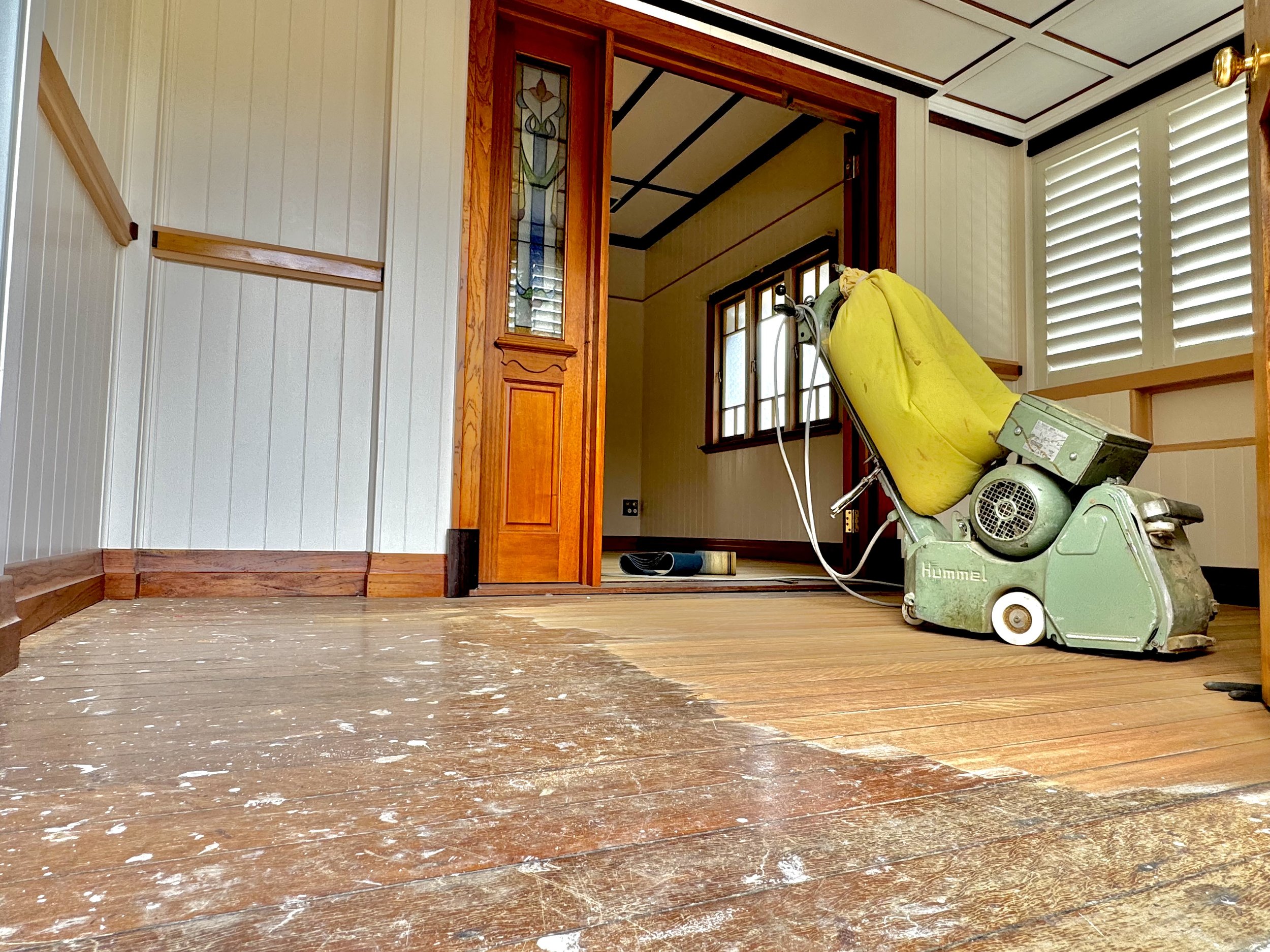 An interior room undergoing renovation with a floor sander machine on the right, a yellow dust bag attached, and scattered dust and wood debris on the floor. The room features wooden trim, stained glass in a doorway, and window shutters.
