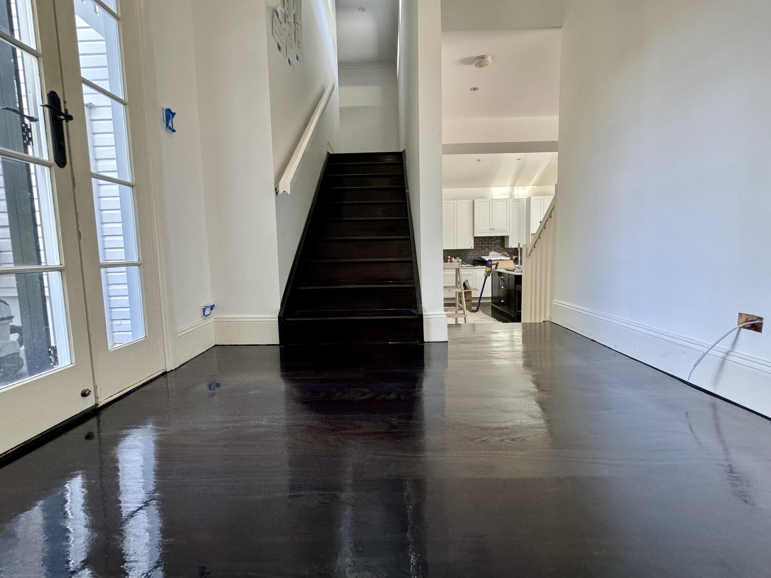 Interior of a house with dark wood flooring, white walls, a staircase, large glass doors, and an open kitchen area in the background.