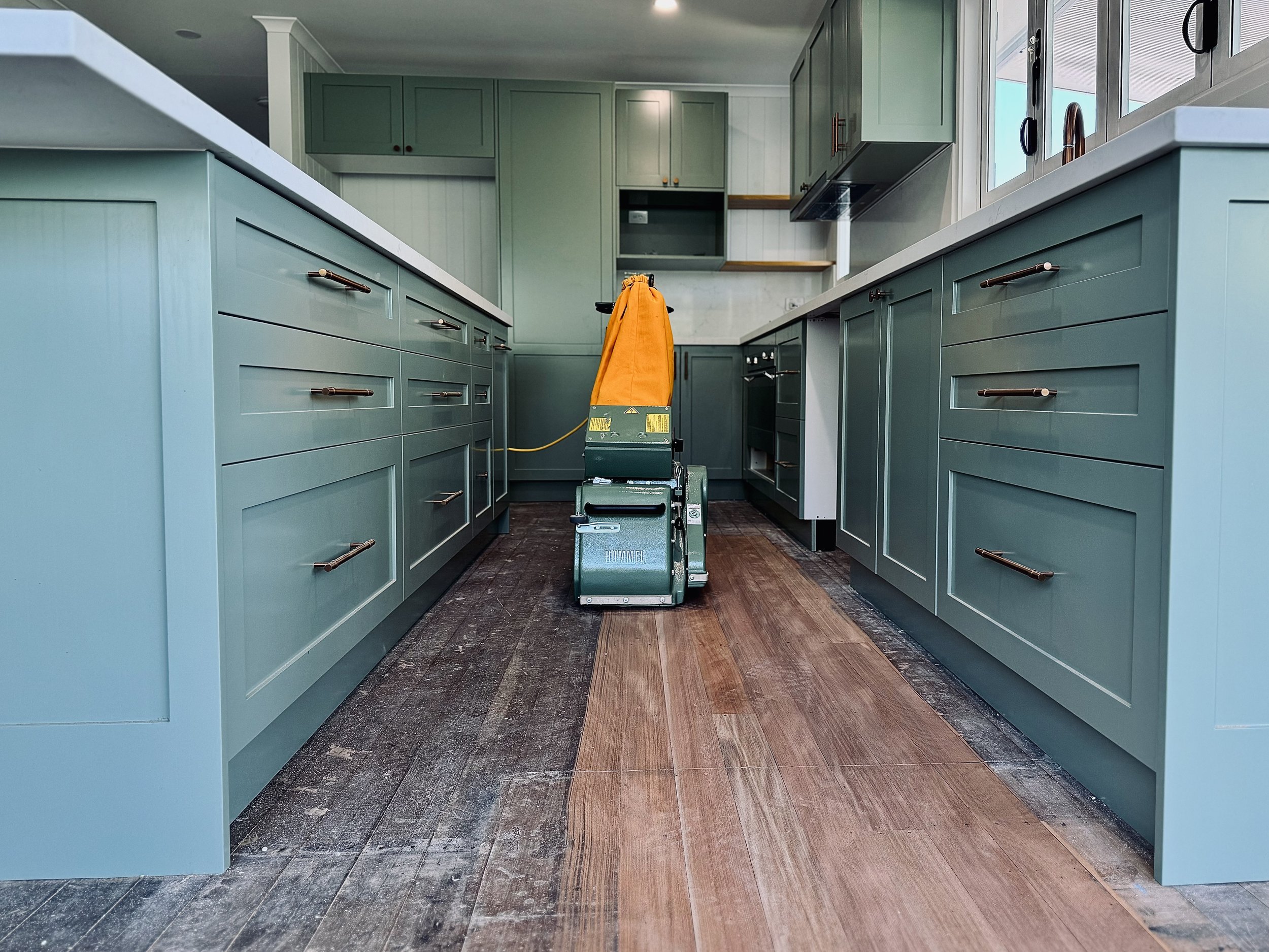 Modern kitchen with green cabinetry and wooden floors, with a floor sander machine in the center.