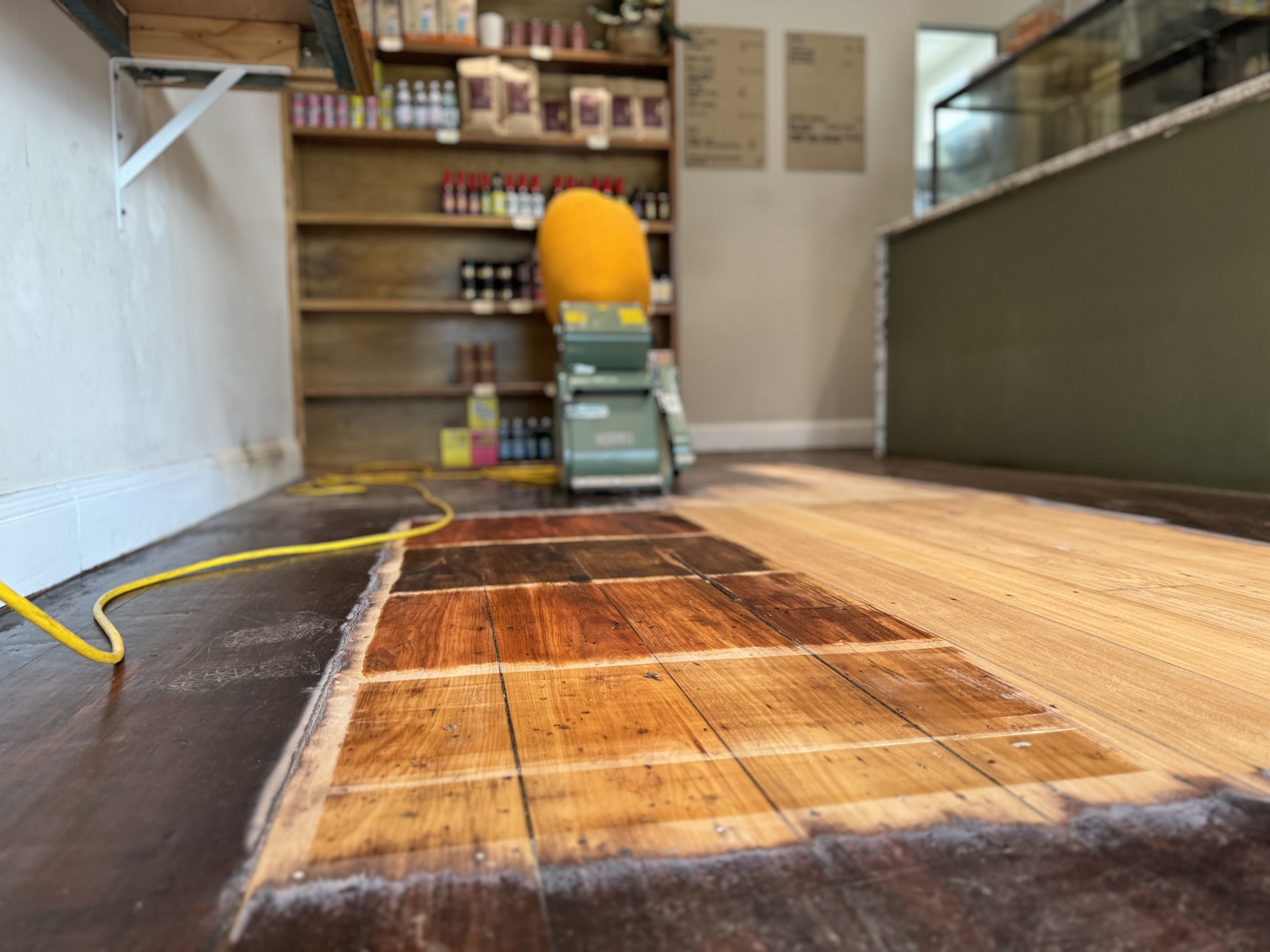Close-up view of wooden flooring being installed or refinished in a shop or cafe setting, with a wall on the left, a shelf with bottles and boxes in the background, and a counter on the right.