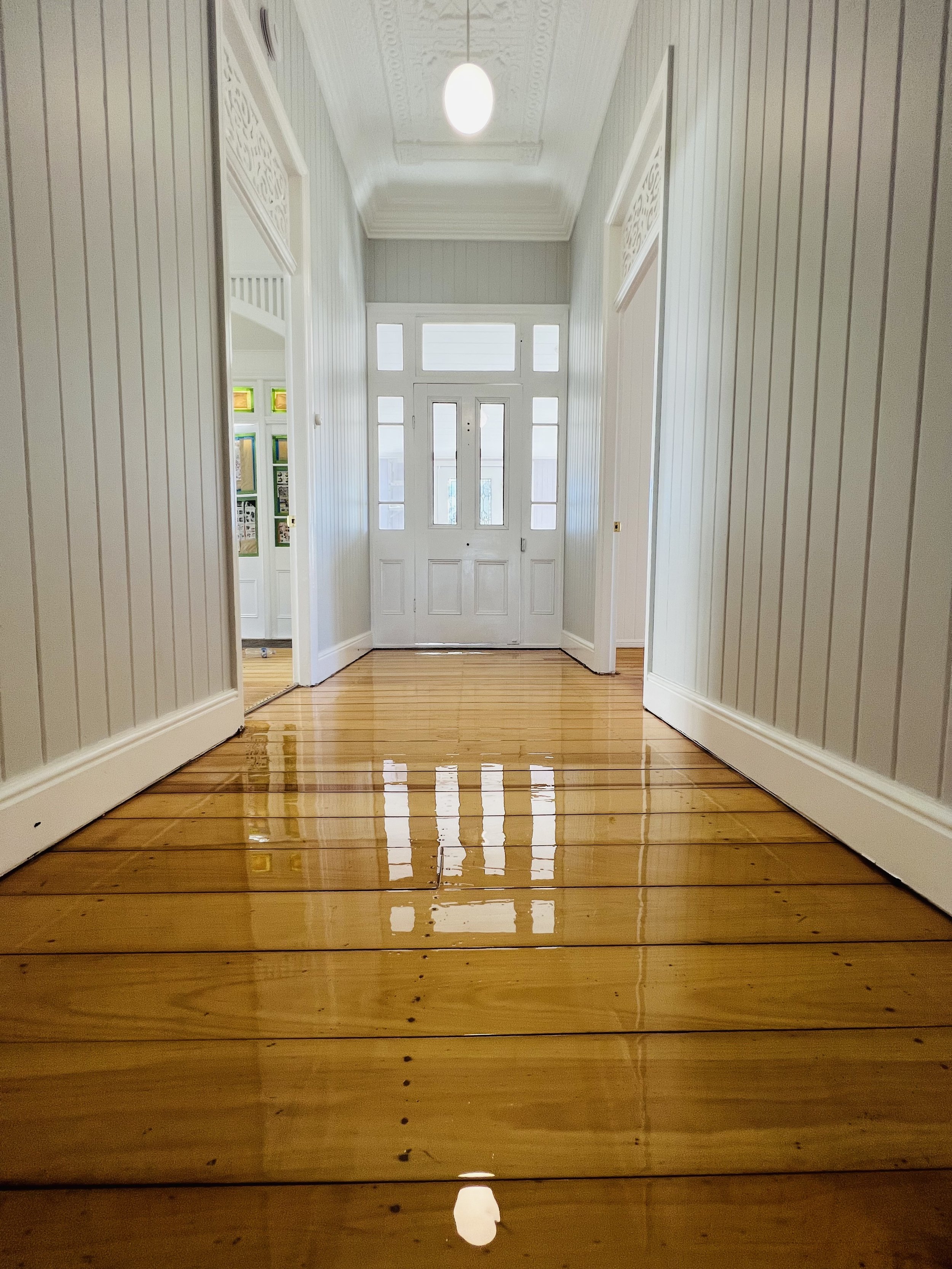 View of a hallway with shiny wooden floors, white paneled walls, decorative crown molding, and a white front door with multiple glass panels, illuminated by ceiling light.